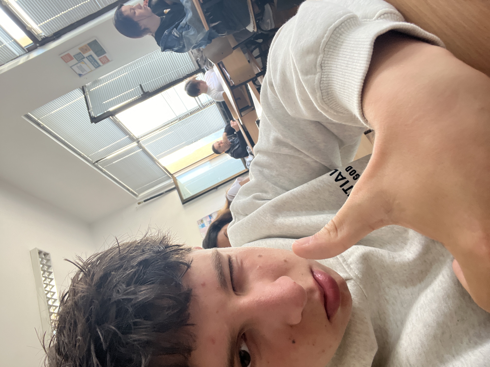 A young male is featured prominently in the foreground of this indoor selfie-style photograph, taken in what appears to be a classroom in Zlatograd, Bulgaria. He is positioned at an angle, looking towards the camera with one eye partially closed in a wink or blink, and a slight smirk on his face. His dark hair is somewhat tousled, and he wears a light gray hooded sweatshirt with partially visible text "NTIAL GOD" (likely "ESSENTIAL GOD" or similar) on the chest area. His right hand is raised, thumb extended, fingers curled, in a casual gesture.
In the background, several other students are visible, seated at wooden desks and chairs, confirming the classroom setting. One individual with dark hair is visible in the upper part of the frame, facing away from the camera, and another in a white top is further back. The room is well-lit by natural light streaming through multiple rectangular windows on the ceiling, each fitted with horizontal blinds. A wall-mounted fluorescent light fixture and a small, colorful poster or display are also visible on the white wall. The overall atmosphere conveyed is casual and relaxed, suggesting a moment of break or lightheartedness during a school day.