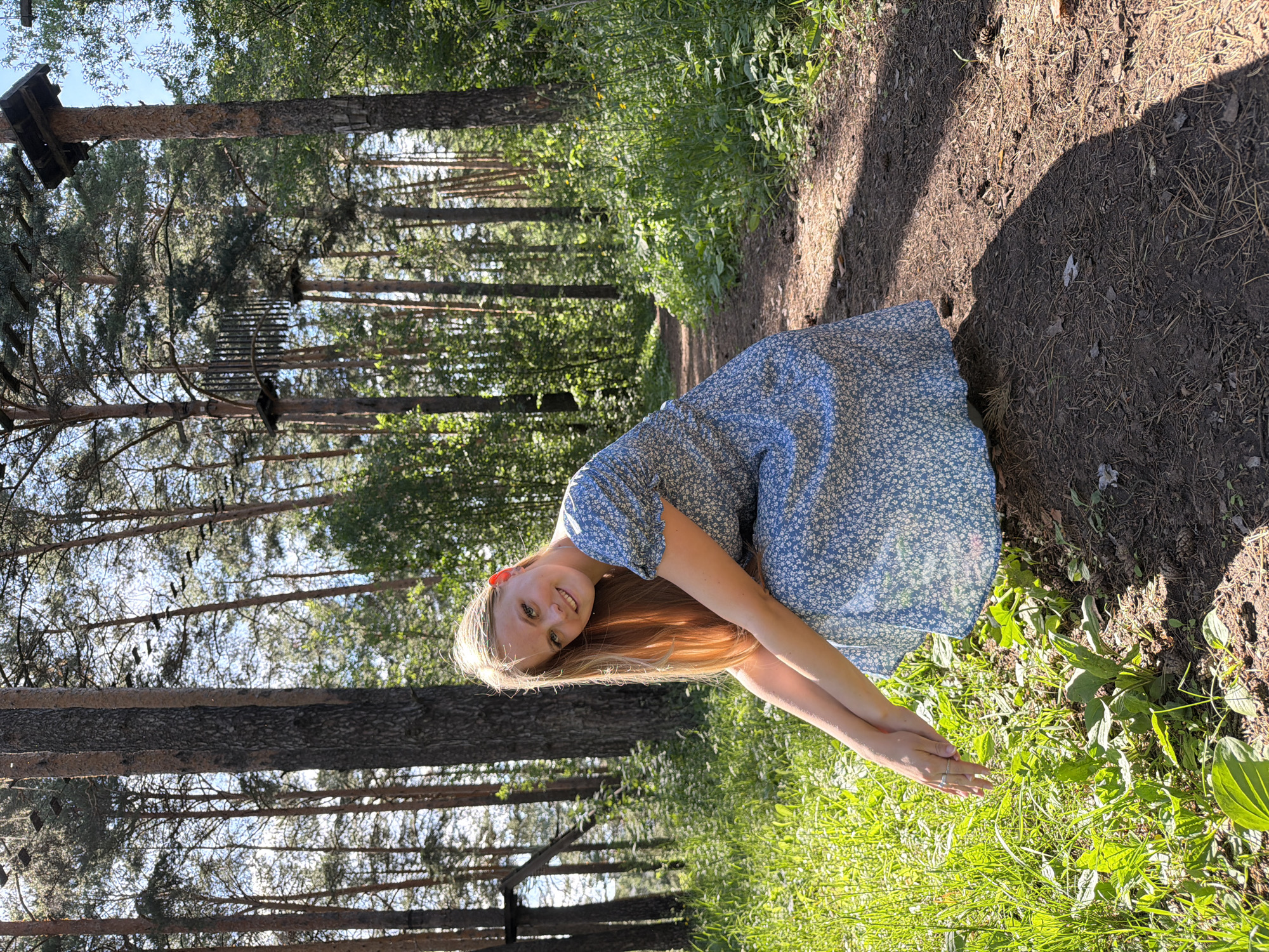 The image shows a young woman with long blonde hair wearing a blue floral dress, crouching on a dirt path in a forest. The path is surrounded by tall pine trees and lush green grass. In the background, there are elements of an adventure park, including wooden platforms and rope bridges suspended between trees. The lighting suggests it is daytime, with sunlight filtering through the trees, casting shadows on the path. The woman is looking at the camera and smiling. The scene appears to be a recreational area within a forested section of Izhevsk, Russia, likely a park or an adventure course designed for outdoor activities.