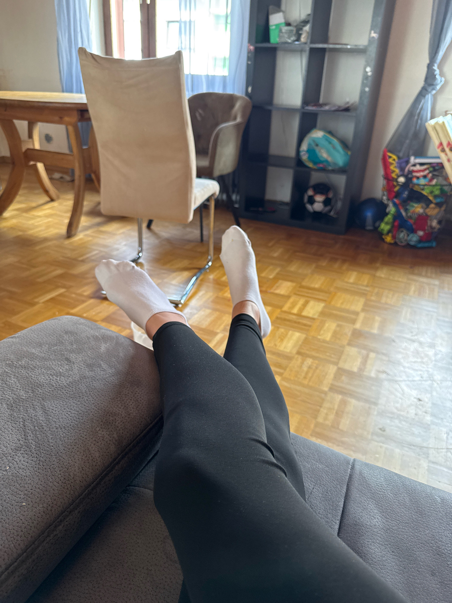 A first-person perspective shot shows a person wearing white socks and black leggings, resting their legs crossed on a dark gray sofa. The legs are positioned towards the right side of the frame. The background features a room with light brown parquet flooring and a large window with sheer white curtains. To the left, a wooden dining table with a light-colored chair is visible. Further back, a dark gray shelf unit stands against the wall, displaying various items including a soccer ball and a turquoise bag. The overall scene depicts a casual indoor setting, possibly a living room or dining area.