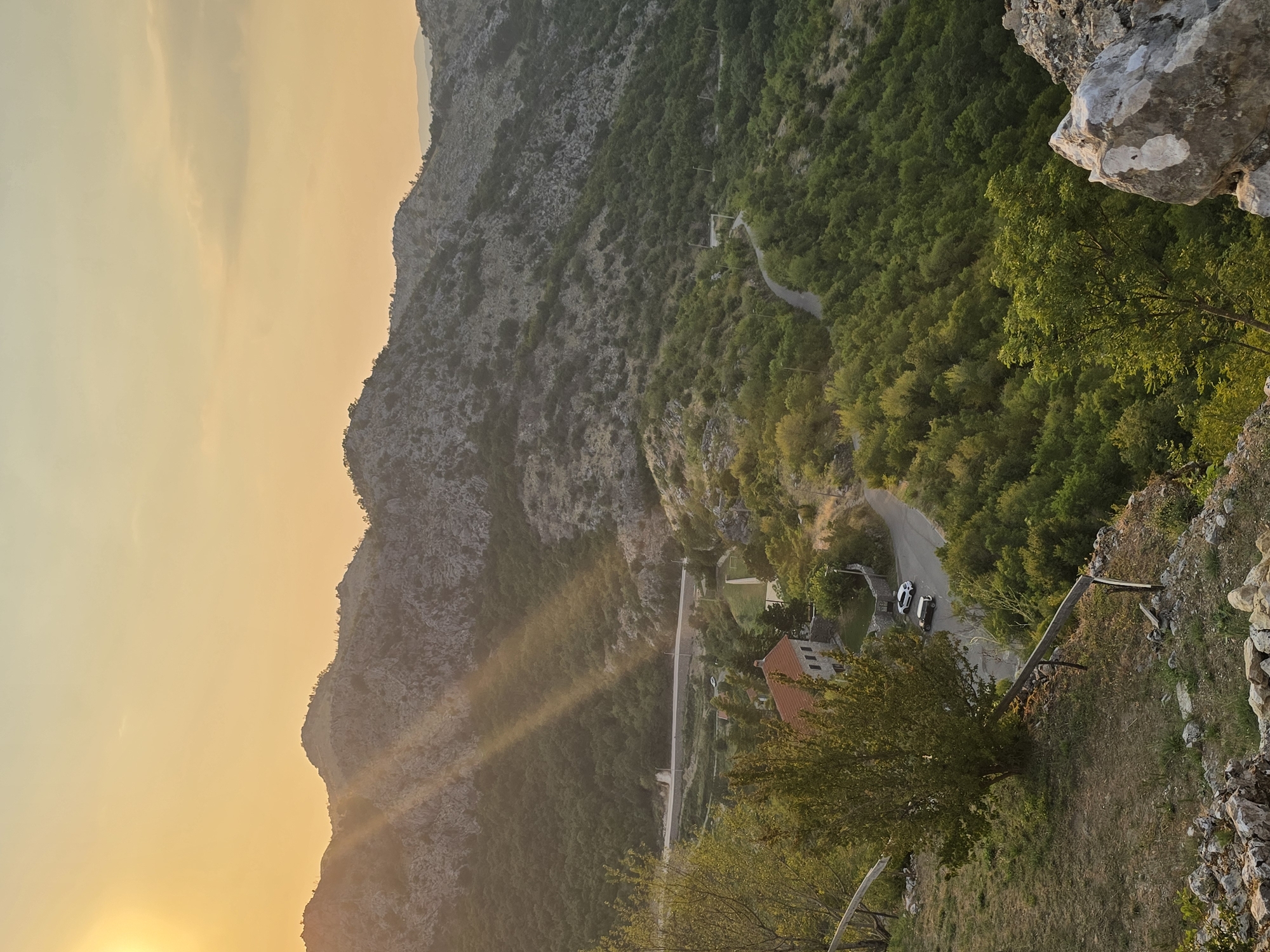 The image captures a scenic view of the city of Medun, Montenegro, during sunset. The warm, golden light of the setting sun casts long shadows across the rugged, rocky mountains that dominate the landscape. A winding road snakes through the lush green hills, dotted with trees, leading to a charming stone house with a red-tiled roof. Two vehicles are parked near the house. The foreground features a grassy slope with a wooden fence and scattered rocks, suggesting a vantage point overlooking the valley. The overall atmosphere is serene and picturesque, highlighting the natural beauty of the Medun area.