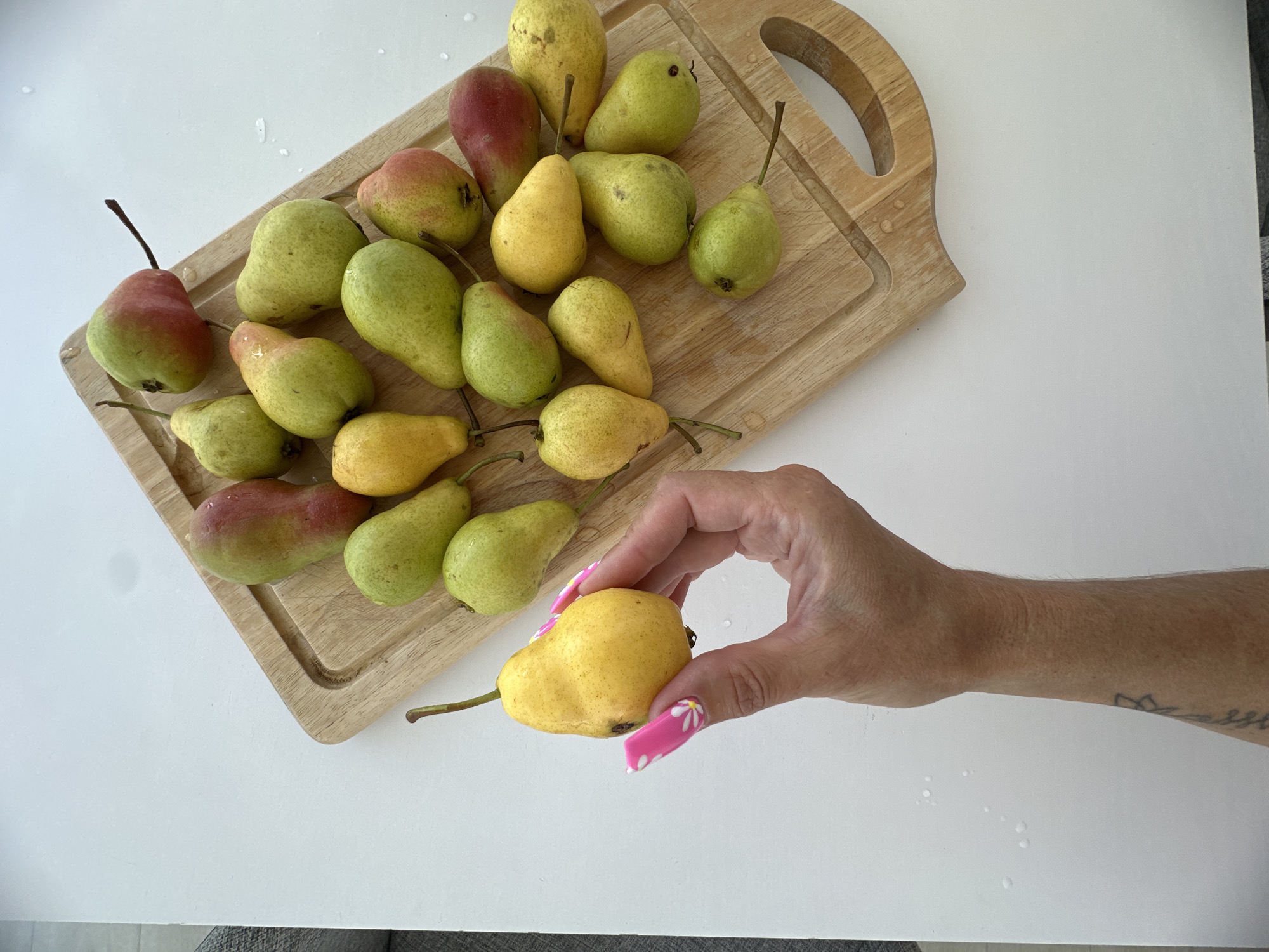 A hand with pink, floral-patterned nails holds a ripe, yellow pear. The pear is plump with a smooth skin and a thin stem. The hand is positioned in the lower-left quadrant of the image, obscuring a portion of a white surface. Behind the hand, a wooden cutting board is laden with a collection of pears. These pears vary in color, ranging from deep red to green and yellow. Some are speckled with red blush. They are arranged in a slightly overlapping manner on the board. The cutting board itself is light-colored wood with a groove around its edge. The entire scene is set against a plain white background, with a few water droplets visible on the white surface to the left of the cutting board. The image does not depict any specific location within the city of Tyumen, Russia.