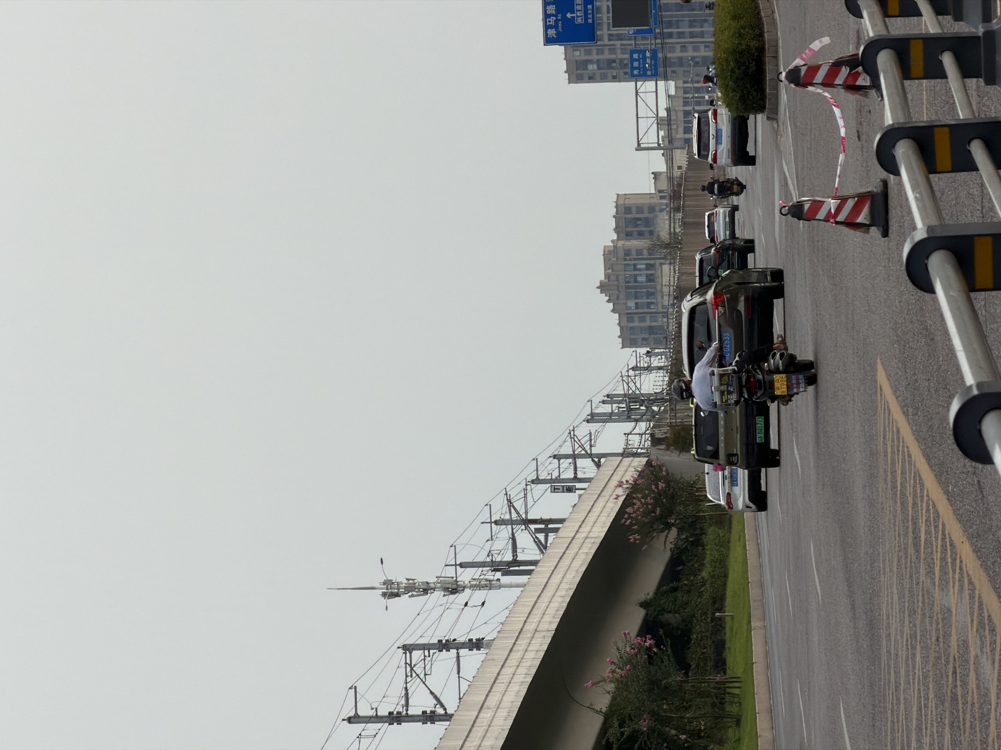 The image captures a street scene in Jiangjin, China, on an overcast day. The foreground features a road with traffic cones and a metal barrier. Several vehicles are on the road, including a motorcycle with a rider, an SUV, and other cars. In the background, an elevated structure, possibly a highway or train track, is visible with supporting poles and overhead wires. Further in the distance, buildings of varying heights, including multi-story structures, line the cityscape. Vegetation, including bushes and trees with pink flowers, borders the elevated structure. A blue road sign with Chinese characters and English text indicating "Jinma Rd" and an arrow pointing upwards is present on the right side of the image. The sky is a uniform light gray, suggesting cloudy weather.