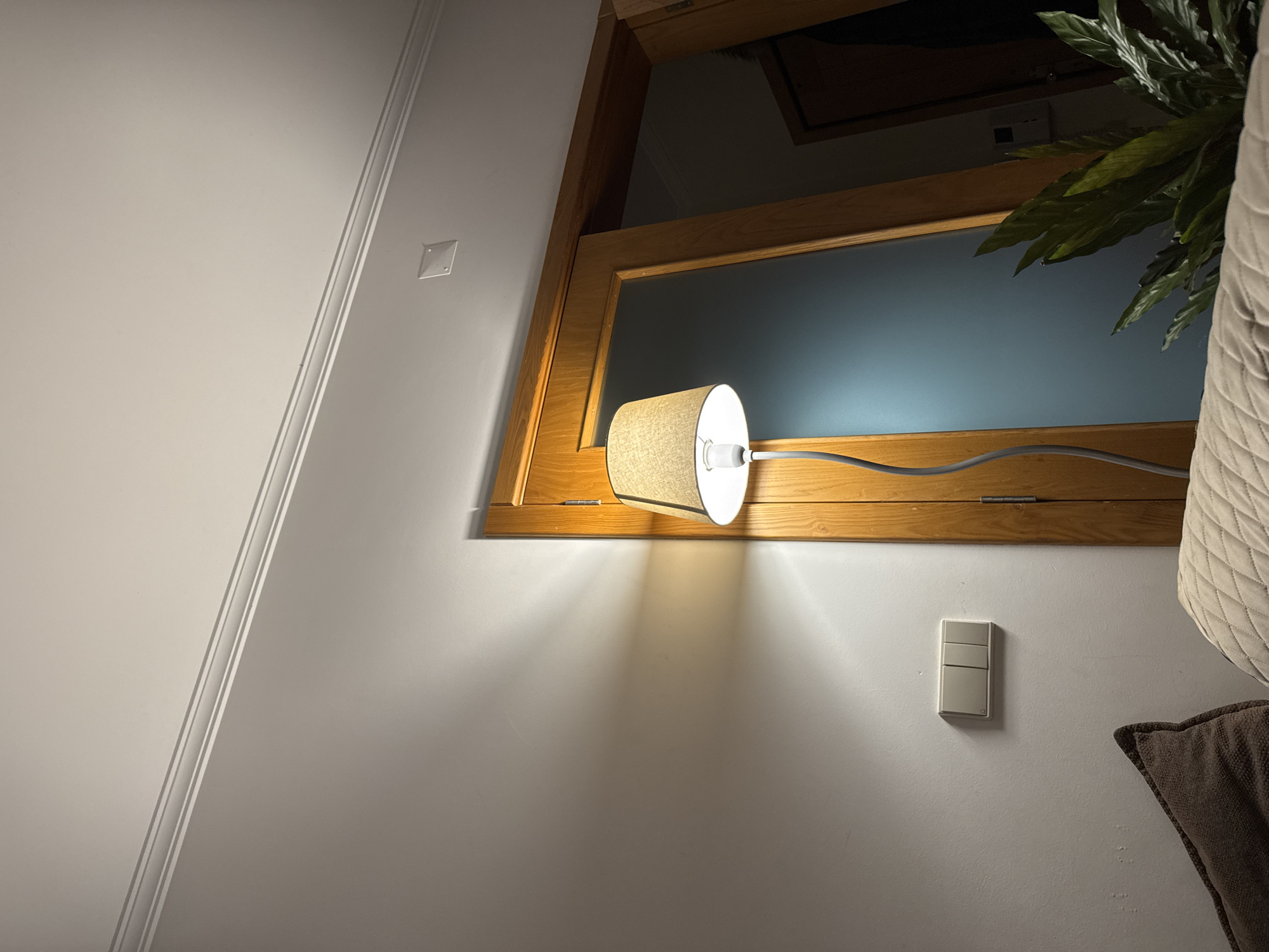 This image depicts an interior scene in Mirandela, Portugal, illuminated by artificial light. A modern floor lamp with a curved grey stand and a beige fabric drum shade is centrally positioned, emitting a warm, yellowish light. The lamp is situated in front of a light-colored wooden door frame, which encloses a door with a frosted, translucent glass panel. To the far right, another dark doorway is visible, revealing a dark interior. A white wall extends from the left to the door frame, featuring a beige three-gang light switch panel mounted horizontally. Shadows radiate from the lamp onto this wall. The white ceiling has decorative crown molding running along its top edge and a small, clear, square-shaped cover near the door frame. In the foreground, the edge of a light-colored, quilted upholstered furniture piece, likely a sofa or bed, is visible, along with a dark brown textured throw pillow on its left. A green, leafy indoor plant is positioned in the lower right, partially obscured by the foreground furniture, its leaves reaching towards the frosted glass door. No individuals are present in the scene.