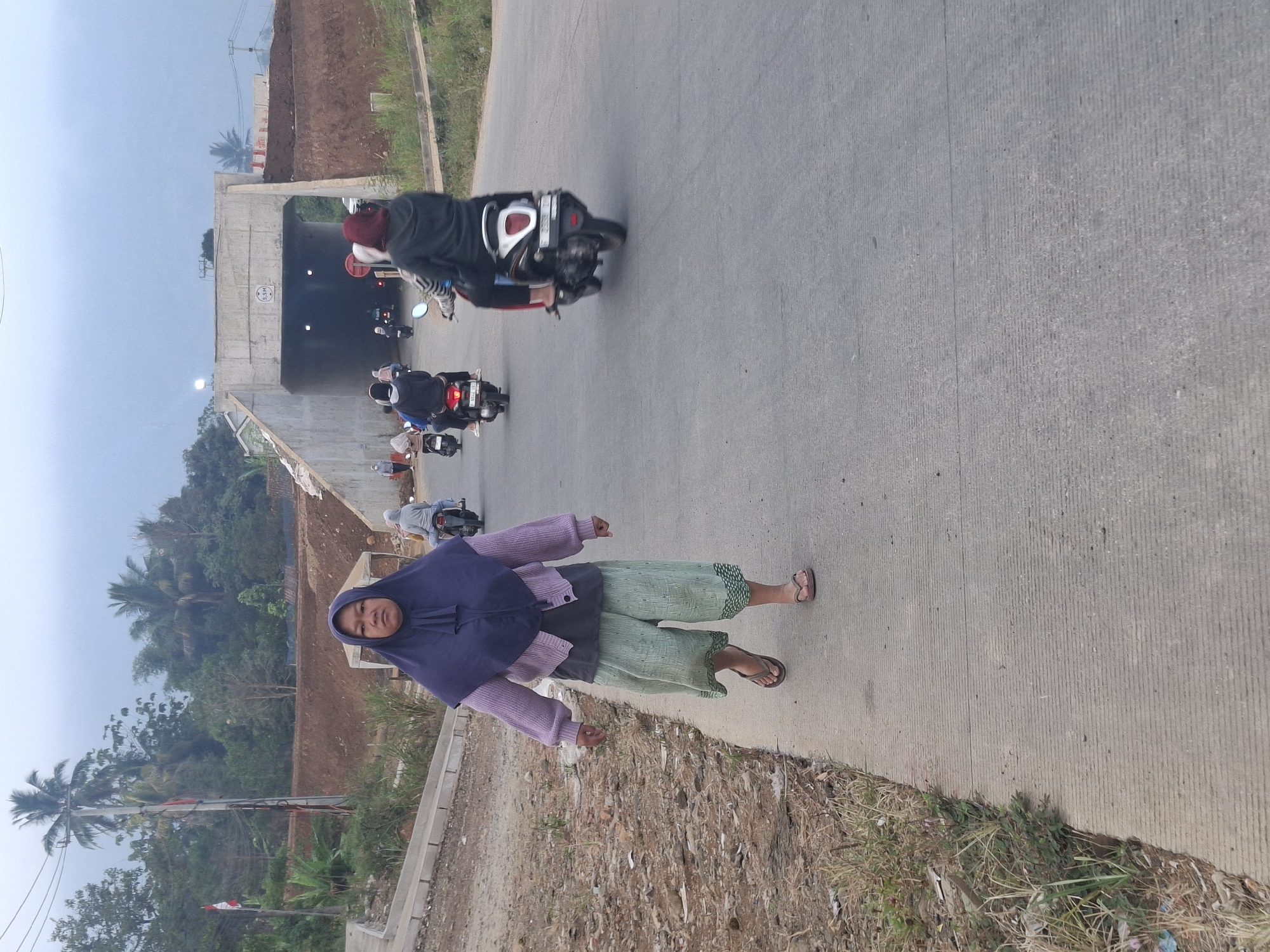 An adult woman walks along a concrete sidewalk on the right side of a paved road. She is wearing a dark purple hijab, a light purple cardigan, a dark gray t-shirt, and loose-fitting light green pants with a patterned hem. She is wearing brown flip-flops. The woman faces forward with a neutral expression. Behind her, several motorcycles are traveling on the road. One motorcycle is directly behind her, ridden by a person wearing a dark gray hooded jacket. Further down the road, other motorcycles are visible. In the background, a concrete overpass or bridge is under construction, with dirt embankments on either side. Trees and foliage line the landscape. The sky is overcast. The scene appears to be in Ciheulang Tonggoh, Indonesia, based on contextual information.