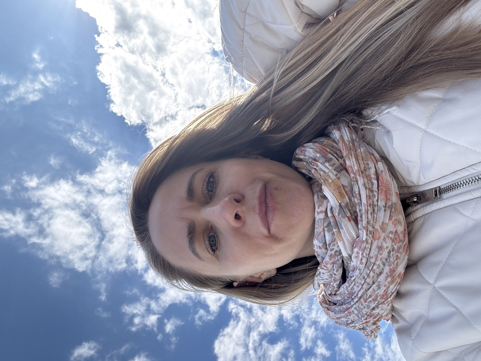 This close-up outdoor selfie, taken in Chaikovsky, Russia, features a woman looking directly upwards at the camera against a bright blue sky with scattered white clouds. Her face occupies the lower-center of the frame, framed by long, straight, light-colored hair, possibly blonde or light brown with highlights. Her eyes are light-colored, appearing blue or grey, and her expression is neutral, perhaps slightly contemplative, with a subtle frown visible between her eyebrows.
She is wearing a light-colored, possibly off-white or beige, quilted jacket, a portion of which is visible on the right side, revealing a silver-toned zipper. Around her neck, she has a light-toned floral scarf with patterns in shades of pink, orange, and muted grey. A small ear piercing can be seen on her left ear. The scene is brightly lit, suggesting a sunny daytime, likely midday or early afternoon, given the intensity of the light and the clear sky conditions. The background consists entirely of the vibrant sky and numerous fluffy white clouds. No text is visible in the image.