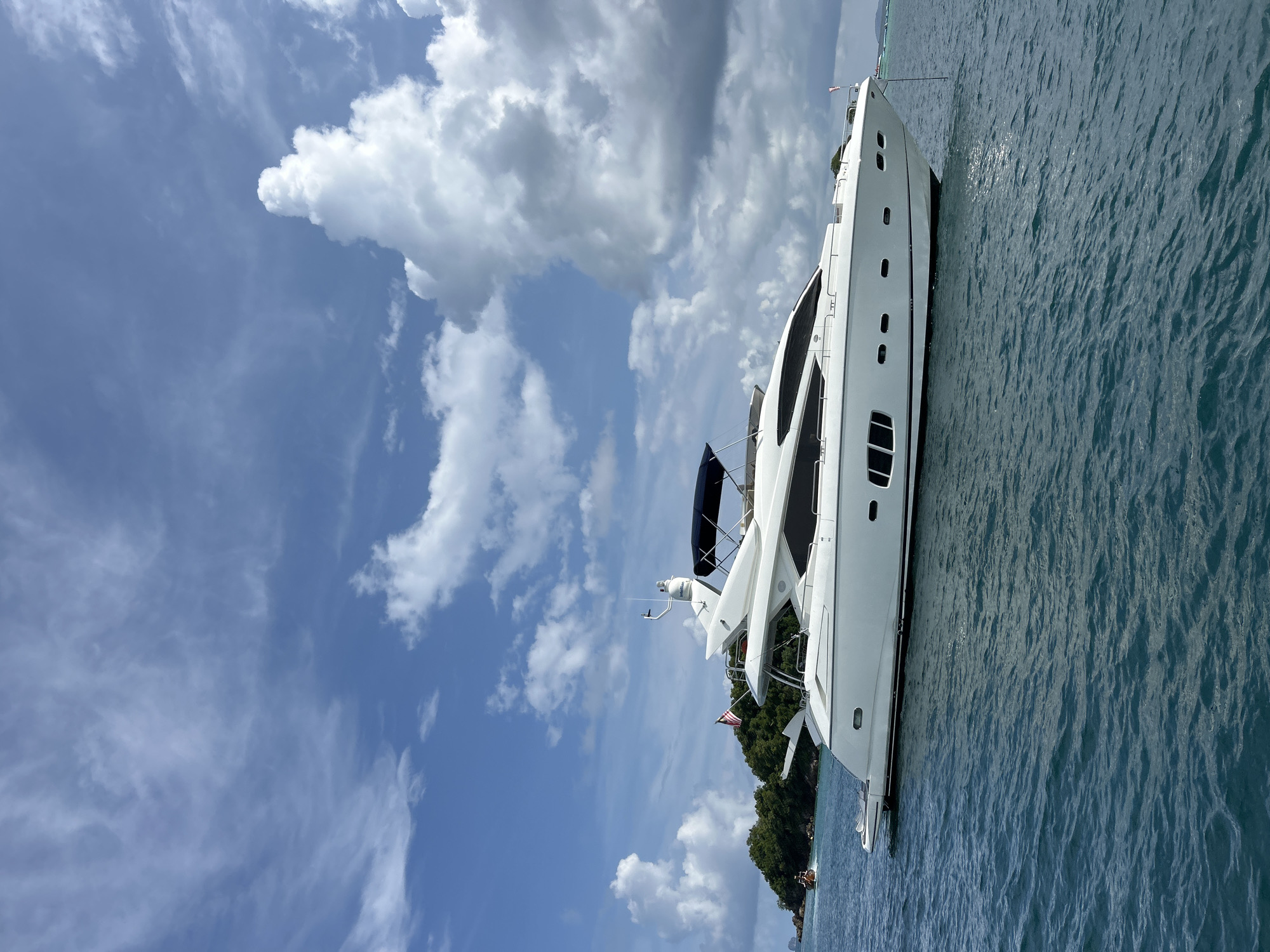 An anchored white luxury yacht is positioned in the turquoise waters of Phru Nai, Thailand. The yacht, a prominent white vessel with dark-tinted windows, is the primary subject, situated in the foreground to the right of the frame. Behind the yacht, a lush green, tree-covered island rises from the sea, partially obscured by the vessel. The sky above is a vibrant blue, dotted with a mix of wispy white cirrus clouds and larger, more voluminous cumulus clouds, suggesting a partly cloudy day. The water exhibits a gentle ripple pattern, reflecting the sunlight. In the distance, to the left of the island, a small orange boat is visible, its presence minimal. The overall scene depicts a serene coastal environment.