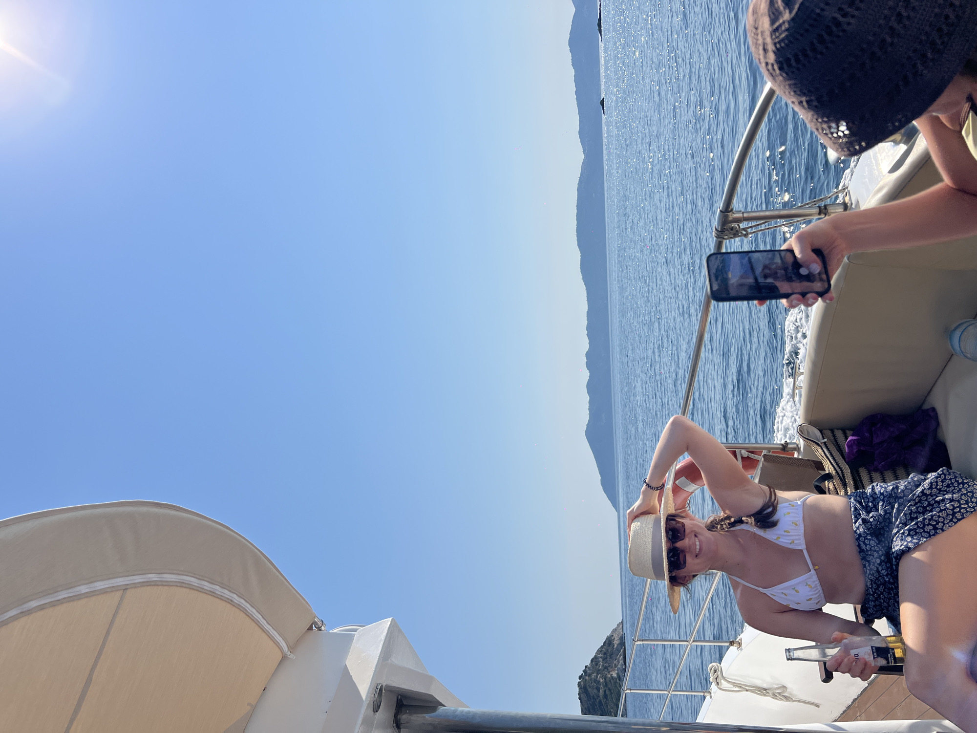 A woman with dark braided hair and sunglasses, wearing a white bikini top and a patterned wrap skirt, sits on a boat with a straw hat on her head. She holds a bottle of beer in her right hand and rests her left arm on the back of her head. Beside her, another person, wearing a brown knitted hat, holds up a smartphone, seemingly taking a photo or video of the woman. The boat is on the sea, with islands in the background under a clear blue sky. The sun is shining brightly. The scene appears to be a leisure activity on the water, possibly in the Aegean Sea near Skiathos, Greece.