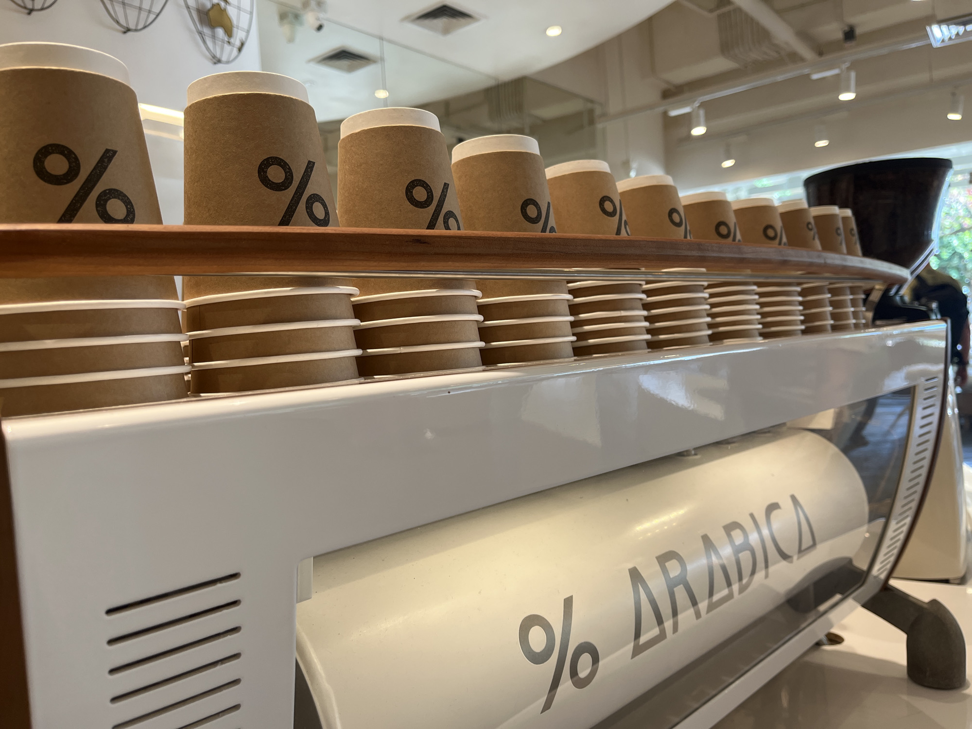 The image shows a row of brown paper coffee cups with a black '%' logo stacked on a wooden shelf above a white espresso machine. The espresso machine has a clear glass front revealing a white cylindrical tank with the text '% ARABICA' in black lettering. The background is blurred, showing a modern interior with ceiling lights and ventilation systems, suggesting a cafe or coffee shop environment. The image was taken in Kuta, Indonesia. There are no people clearly visible in the foreground.