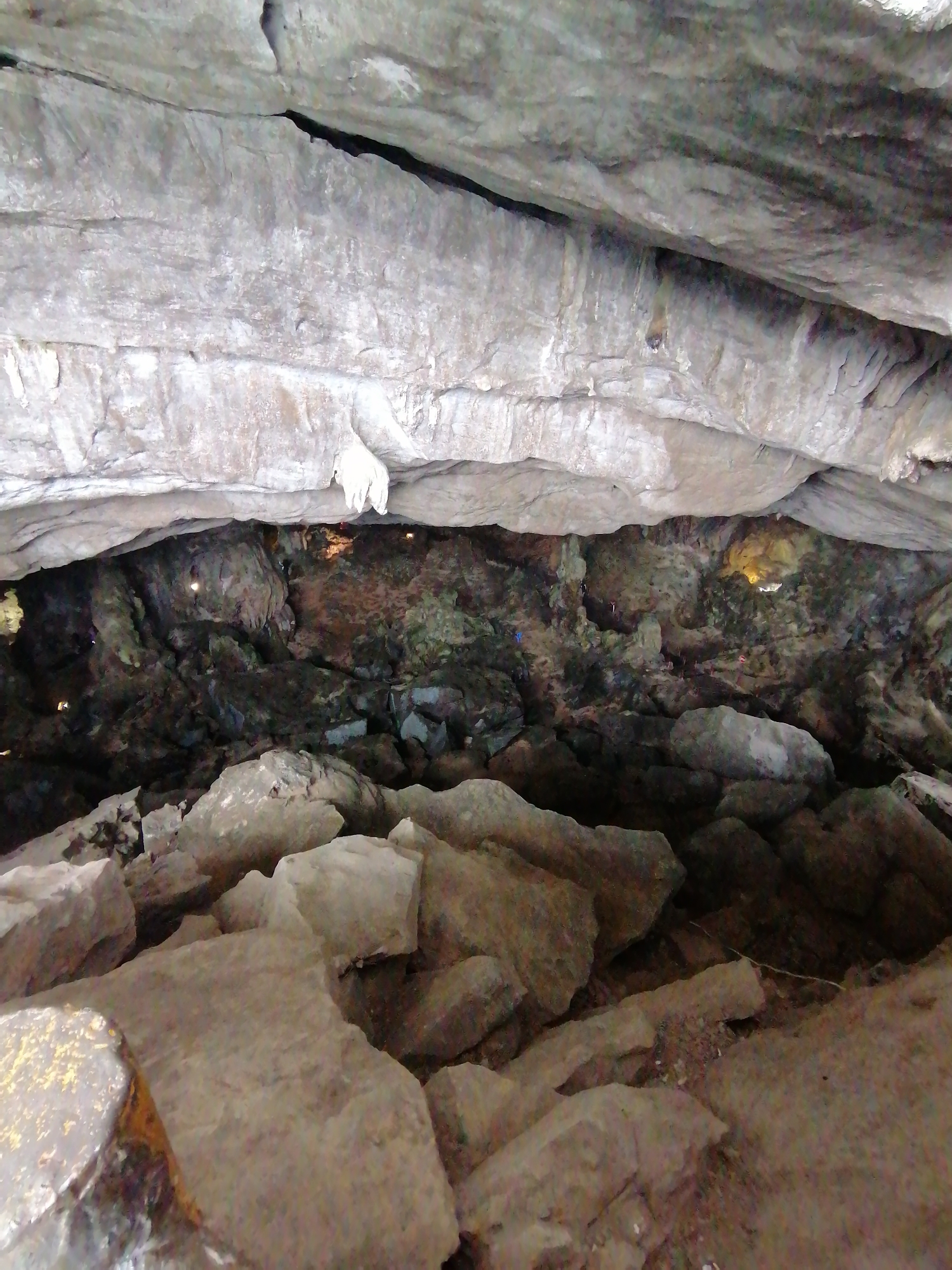 The image presents a view from within a natural cave or a large rock overhang, characterized by massive, textured rock formations. The foreground and mid-ground are dominated by a low, sprawling cave ceiling and walls made of light grey and brownish-grey rock, exhibiting visible strata and erosion patterns. A prominent white, finger-like rock formation, possibly a stalactite, hangs from the ceiling on the upper right side.
Below this rock canopy, the scene transitions into a darker, recessed chamber or passageway of the cave. The walls and floor of this deeper section are composed of rough, uneven rock surfaces, appearing dark brown, black, and subtly green in places, suggesting moss or mineral deposits. Scattered throughout this deeper area are several small, distinct points of artificial light, illuminating specific features on the cave walls and hinting at visitor infrastructure.
The ground directly in front of the viewer is covered with large, angular boulders and fragmented rocks, creating a rugged and uneven terrain. No human activity or visible text is present in the frame. The overall impression is one of natural grandeur and a dimly lit, ancient geological environment, consistent with a cave system found in karst regions like Bai Chay Ward, Vietnam.