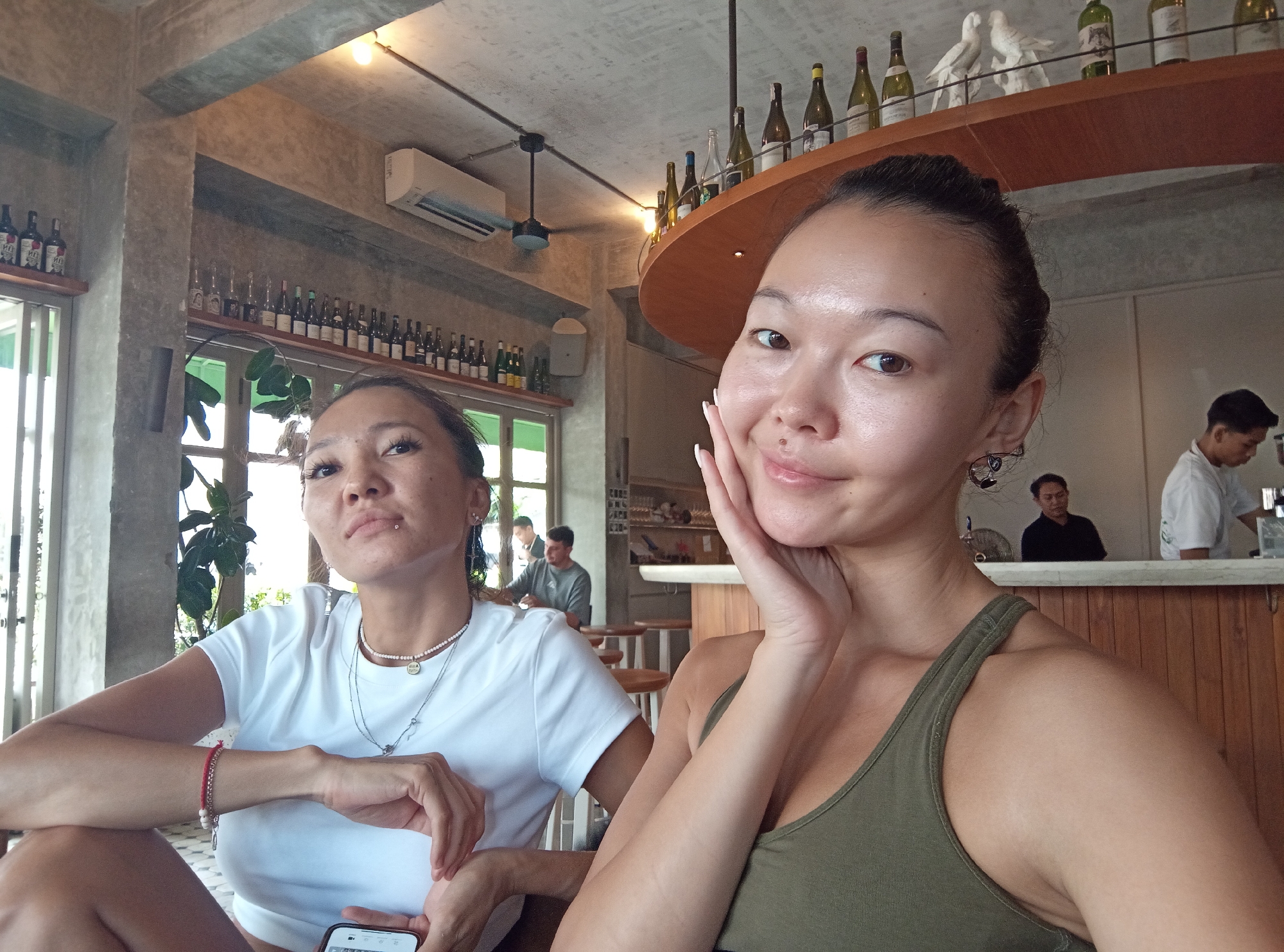 Two women are seated at a table inside a building in Mengwi, Indonesia. The woman in the foreground wears a green tank top and has her hand on her cheek. The second woman wears a white t-shirt, jewelry and holds a mobile phone. Both women have their hair pulled back. The space appears to be a café, with a bar in the background staffed by a man in a white shirt and other patrons at tables. Bottles of wine are displayed on shelves around the room, and decorative items are placed on top of a bar shelf. The interior has a somewhat industrial feel with concrete and wood elements.