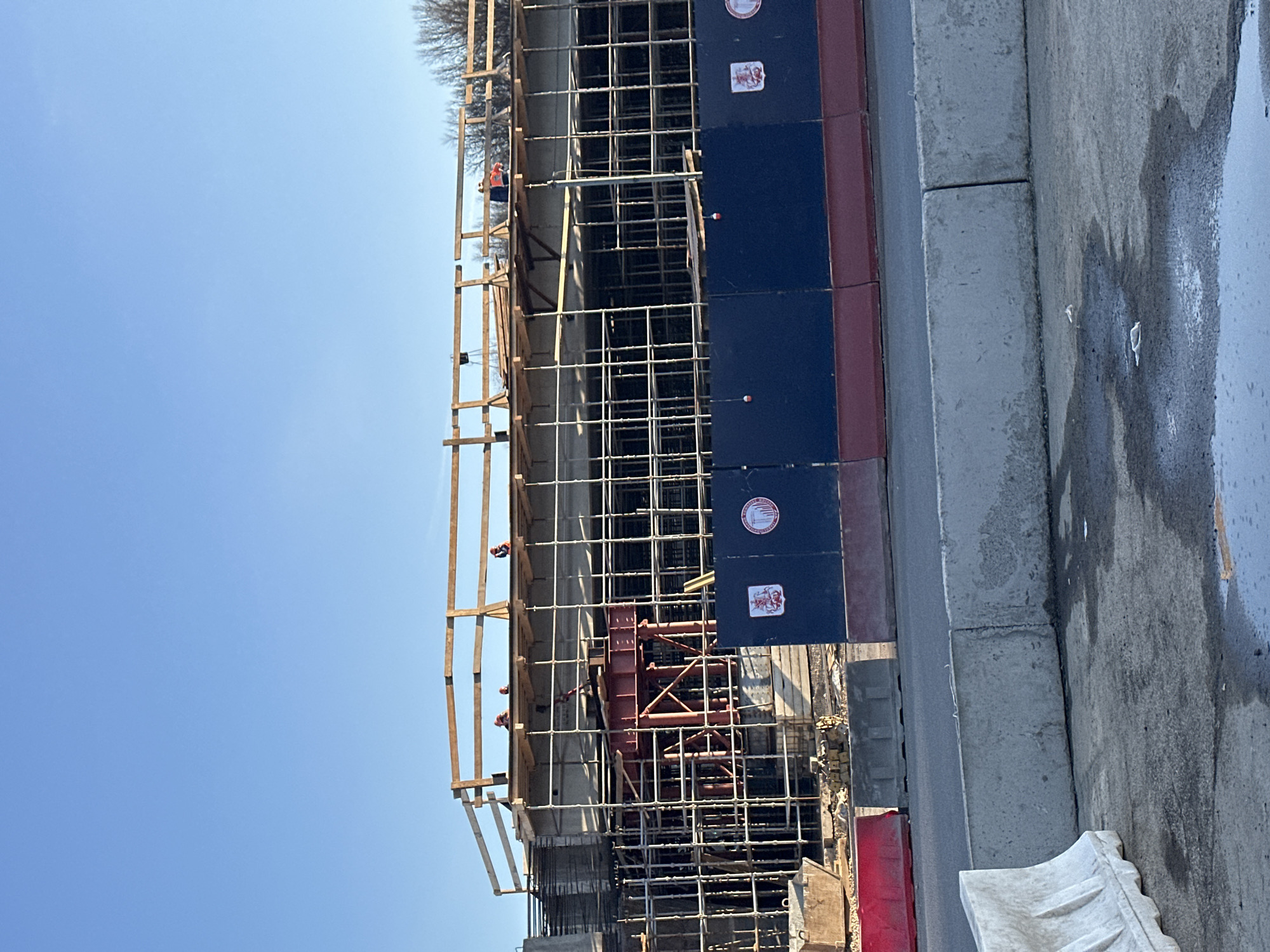 The image depicts a construction site in Moscow, Russia. The focus is on the scaffolding and framework erected for a new building. Several construction workers wearing orange vests are visible on the scaffolding. Construction panels in dark blue and red are incorporated into the structure, bearing round emblems with Russian text and imagery. Below the construction site is a gray concrete curb along a road, with water stains on the surface. In the background, the sky is clear and blue, with bare tree branches visible on the horizon.