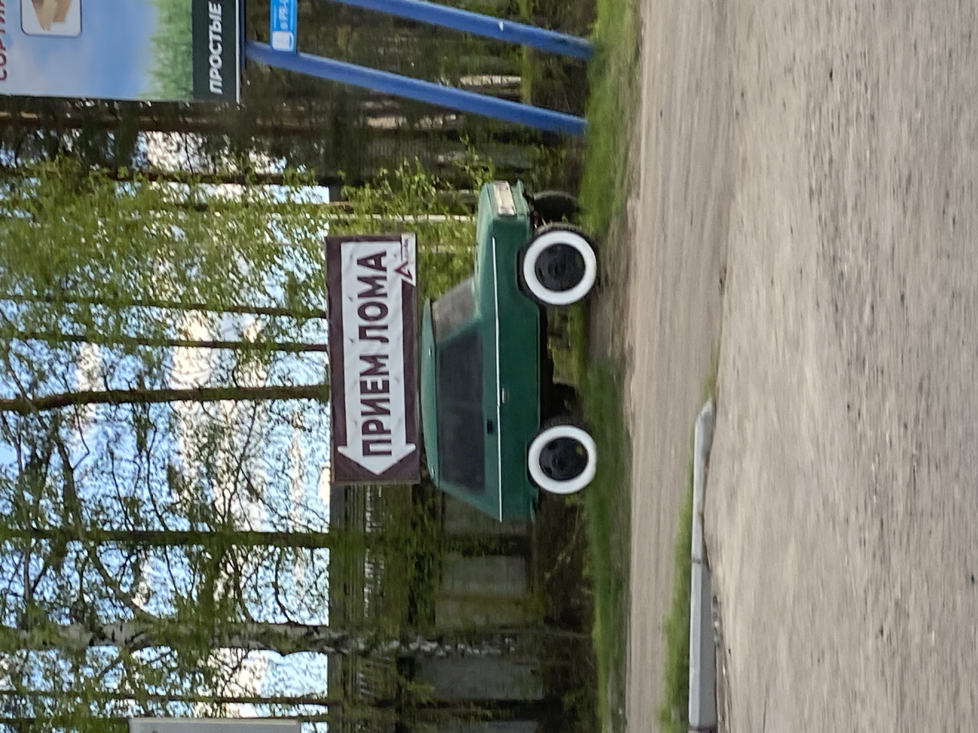 A green automobile with white-wall tires stands perpendicular to the edge of a concrete or asphalt road. A sign with Russian text "ПРИЕМ ЛОМА" is mounted above the car. The location appears to be a rural area near Chaikovsky, Russia, as the background includes trees and shrubs. Another sign, with the text "СОРТИР...", is visible in the upper left, suggesting a commercial or industrial area.
