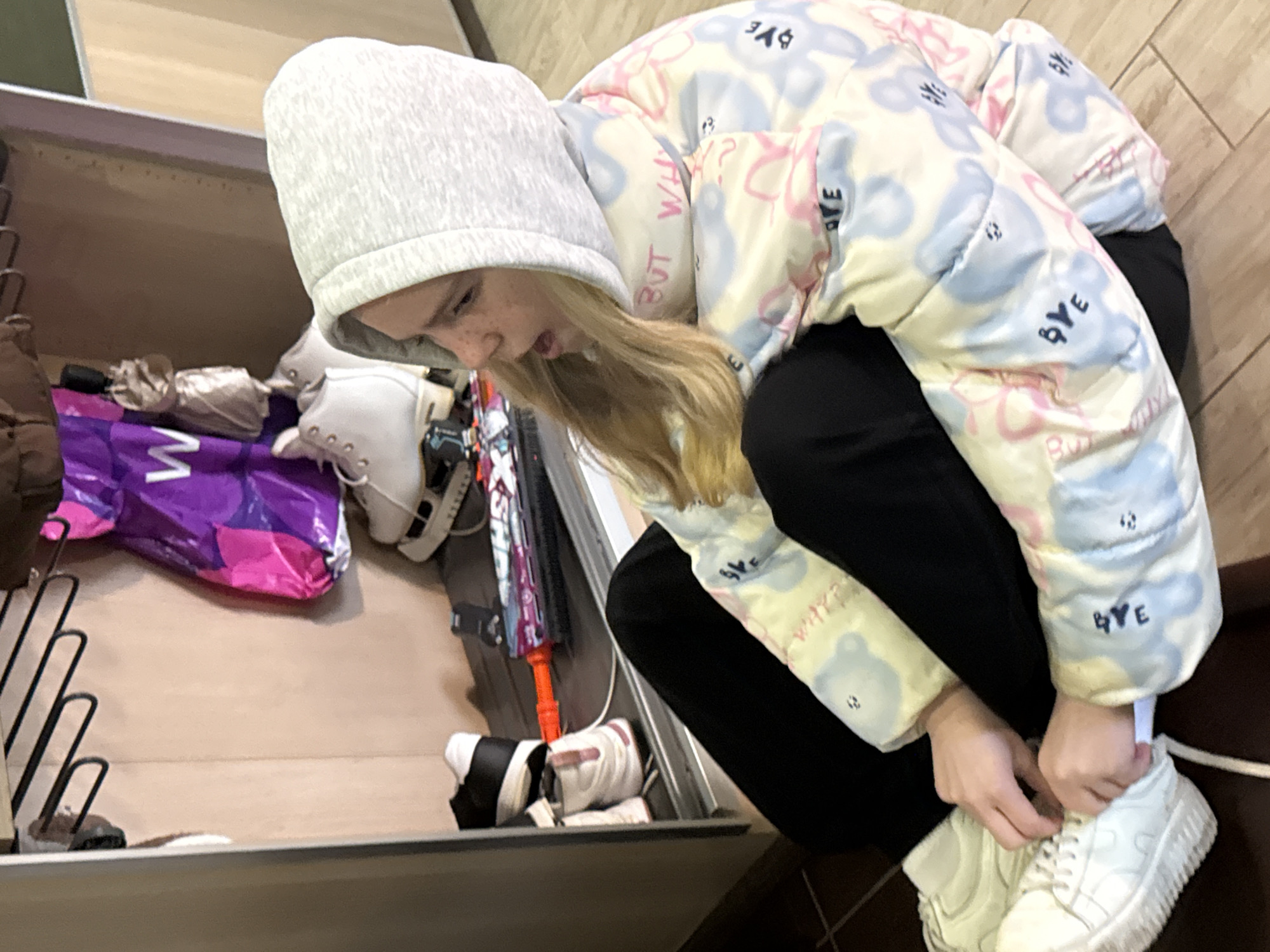 A young person wearing a gray hooded sweatshirt and a light-colored patterned jacket is crouching and tying the laces of a white sneaker. They are indoors, next to a shelf or drawer containing various items, including shoes, a shopping bag, and an umbrella. The location is not discernible from the image.