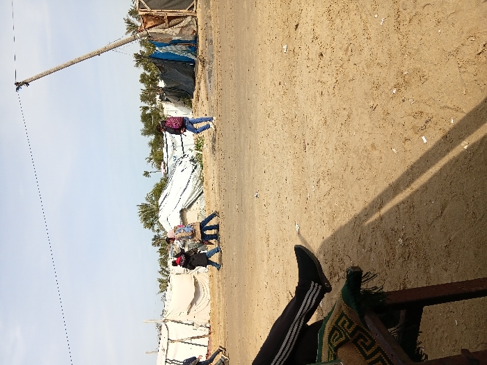 A sandy area shows makeshift shelters and utility poles. People walk along a dirt path. A person wearing dark pants with white stripes and black shoes rests their legs.