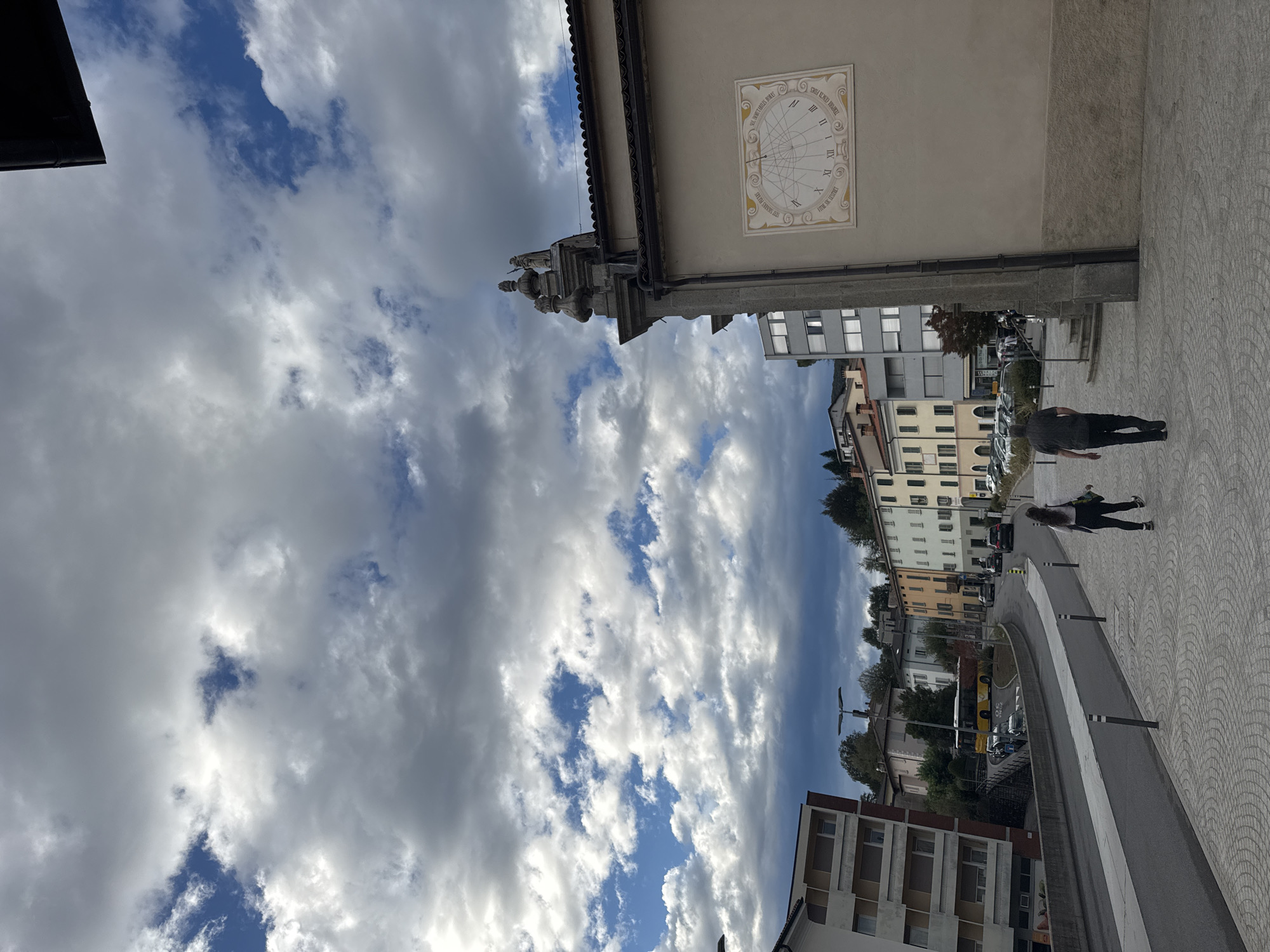 In Balerna, Switzerland, a street scene unfolds under a dramatic sky filled with dark, cumulus clouds interspersed with patches of blue. The foreground features a patterned stone walkway where two individuals are walking away from the camera. A woman, on the left, wears dark leggings and a light-colored top, carrying what appears to be a skateboard. To her right, a man is dressed in dark trousers and a dark short-sleeved shirt.
The street curves to the left, lined with a variety of buildings. On the immediate left, a modern multi-story building with balconies and light-colored facades dominates the view. Further down the street, a bright yellow bus is visible, along with parked cars and other vehicles. The buildings on the opposite side of the street showcase a range of architectural styles and colors, from light yellow and cream to muted pastels. Trees are scattered throughout the urban landscape.
On the right side of the image, a building with a light-colored facade displays a large, circular sundial. The sundial has Roman numerals and intricate markings. Above the sundial, architectural elements, including decorative finials and statues, adorn the building's exterior, suggesting it might be a historical or religious structure. The overall impression is of a partially cloudy day in a European town, with a mix of modern and older buildings.