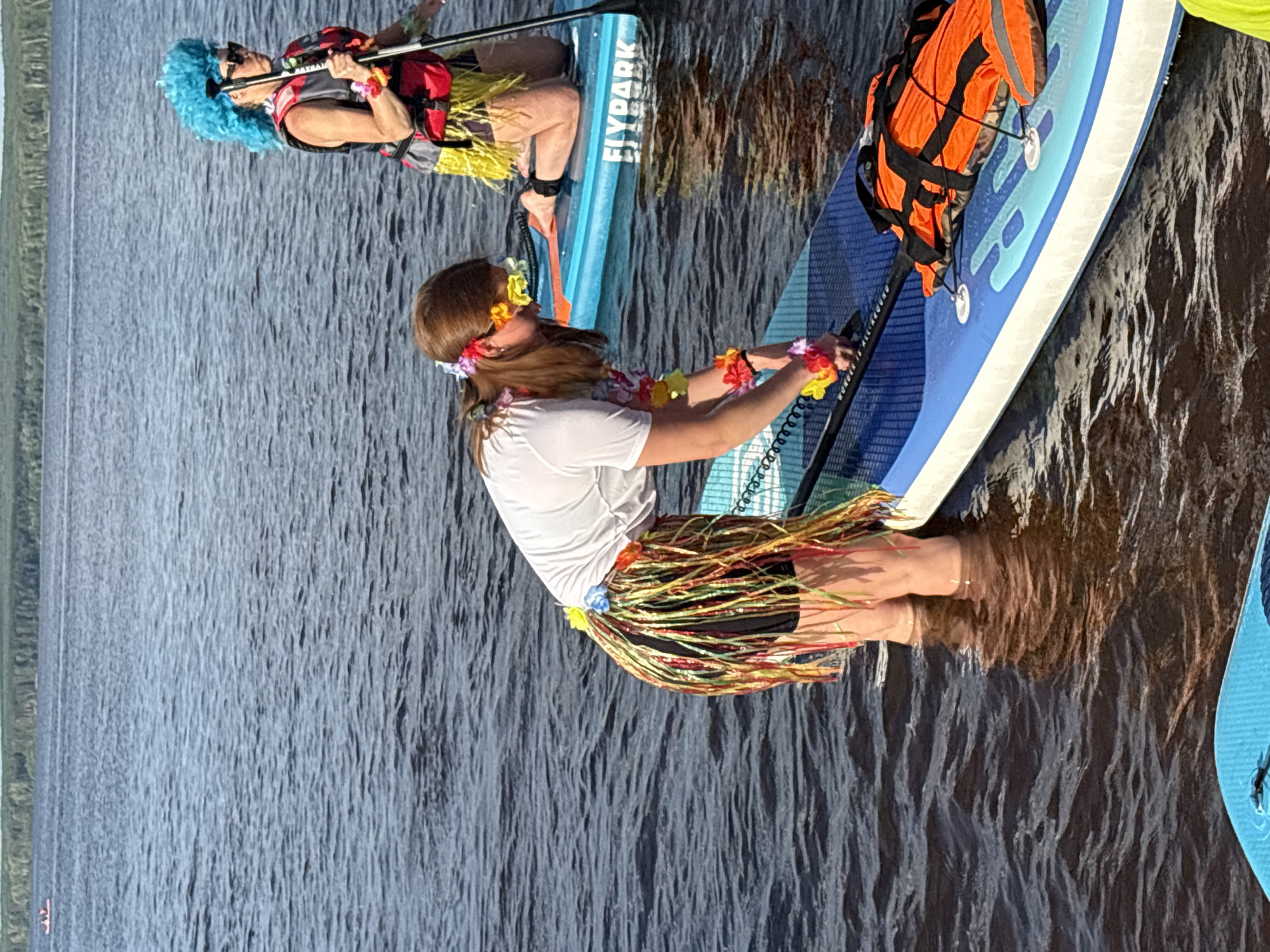 Two individuals are paddleboarding on a body of water, likely a river or lake. The person in the foreground, with reddish-brown hair, is wearing a white t-shirt, black shorts, and a colorful grass skirt with flower accessories around her neck, wrists, and hair. She is bent over her paddleboard, appearing to adjust something. Her legs are submerged in the water up to her knees. The paddleboard is blue and white with the word "FLYPARK" visible. An orange life vest is secured to the front of the board.
The second individual, in the background, has bright blue, spiky hair and is wearing a red and grey life vest over a yellow grass skirt. She is kneeling on her paddleboard and holding a paddle. The environment appears to be outdoors, with a treeline visible in the distance and a calm, dark body of water. The sky is overcast. The location is not identifiable as Chaikovsky, Russia based on the provided image.
