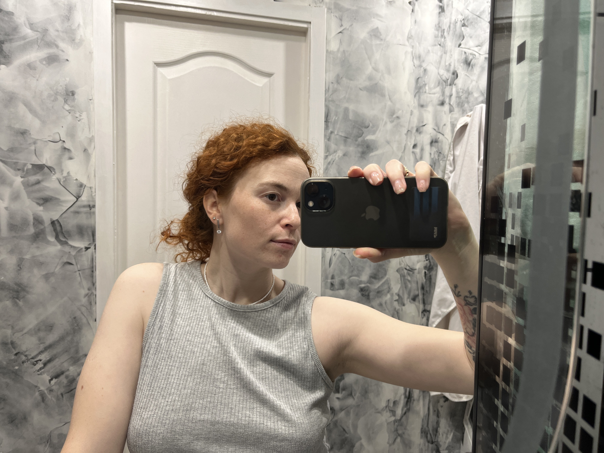 A woman with red, curly hair takes a mirror selfie indoors, holding a dark-colored smartphone with a visible Apple logo in her right hand. She is looking slightly to her right with a neutral expression. She wears a light grey, ribbed tank top, small dangling earrings, and a light-colored beaded necklace. Her fingernails have a light polish.
The background shows a white door with a curved panel design on the left side of the frame. To the left of the door and behind her, the wall features a distinctive grey and white marbled or textured pattern. In the reflection on the far right, elements of the room are visible, including a dark, grid-like pattern, possibly from a shower screen or window covering, and a white garment or towel hanging in the background. A tattoo with what appears to be a floral or ornate design is visible on her left forearm, partly obscured by the phone and reflections. The image captures an everyday moment in what appears to be a private residence in Chaikovsky, Russia.