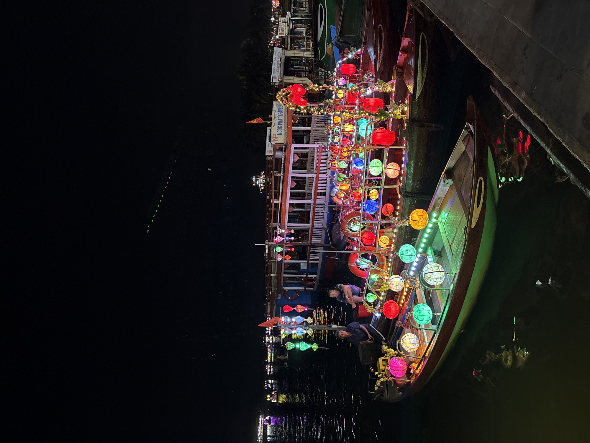 The image captures a night scene in Hội An Ward, Vietnam, featuring boats adorned with colorful lanterns moored along a waterway. The boats are decorated with strings of globe-shaped lanterns in various colors, including red, green, yellow, and blue, illuminating the scene. A larger boat is visible with multiple levels. People are seen aboard one of the boats. The water reflects the lights, creating a shimmering effect. The background shows buildings and other lights along the shore.