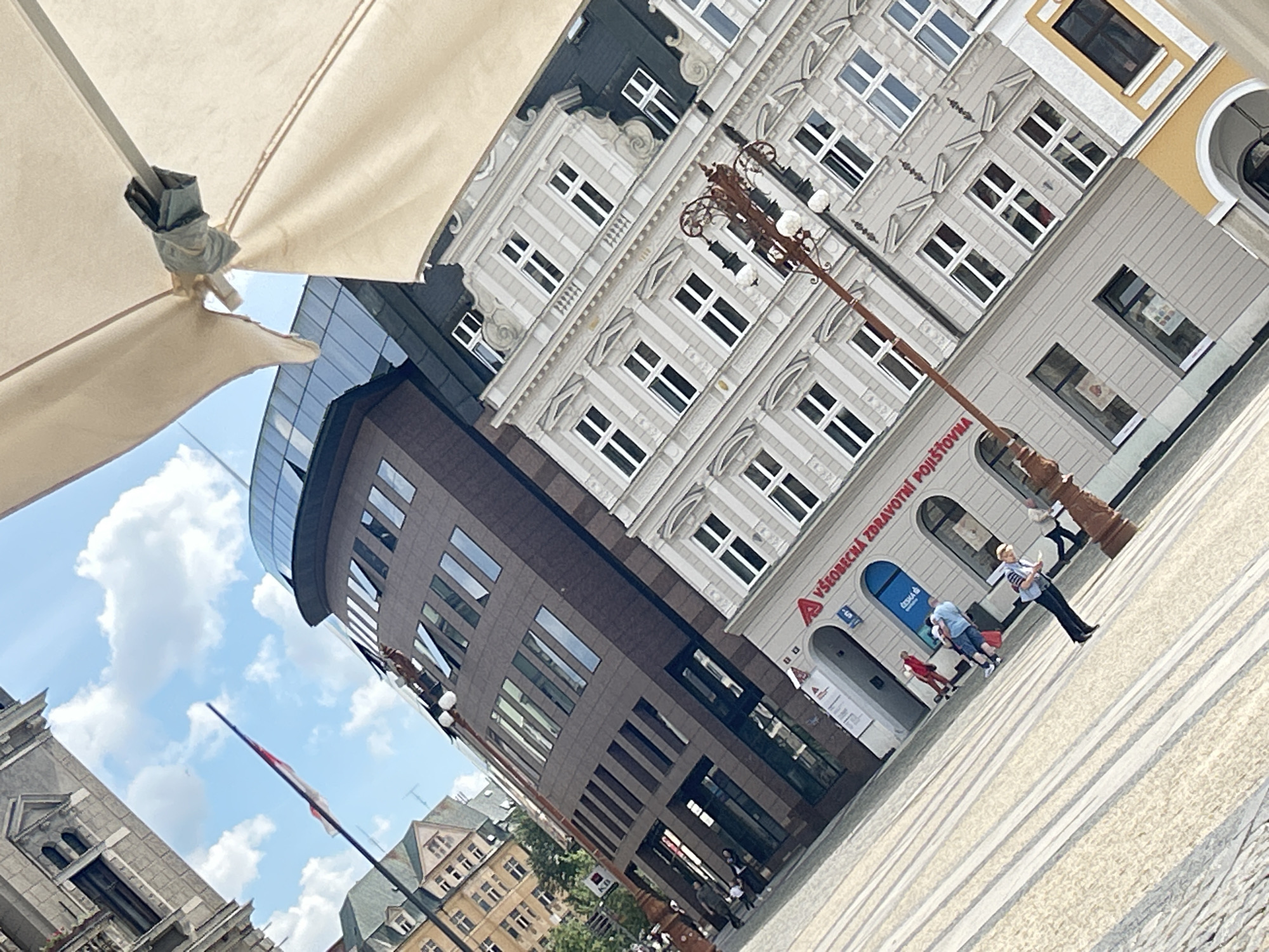 The image presents a tilted, dynamic view of an urban square or street, blending historical and modern architecture under a bright, partly cloudy sky. A large, beige outdoor umbrella or awning occupies the upper left foreground, partially framing the scene.
On the right, a multi-story building features a light gray, classically styled facade with intricate architectural details, including decorative window surrounds and cornices. It has numerous evenly spaced rectangular windows with white frames. The ground floor of this building houses a business with a blue arched entrance and displays the prominent text "VŠEOBECNÁ ZDRAVOTNÍ POJIŠŤOVNA" (General Health Insurance Company) and a visible "A" logo, along with partial "ČESKÁ" text, confirming a Czech location.
To the left, a modern, curved building rises with dark brown or gray paneled walls and large, tinted rectangular windows. Its top section features a glass curtain wall. Further in the background, glimpses of other traditional buildings with gabled roofs are visible.
An ornate, dark-colored lamppost with multiple globes stands prominently in the mid-ground, extending diagonally from the lower right. The ground is paved with light-colored stones, laid in a linear pattern, indicating a pedestrian area.
Several people are visible in the square. A woman with blonde hair, dressed in a striped top and dark pants, walks across the paved area. A small group, including a man in a blue shirt and another person in red, gathers near the entrance of the health insurance building.
The sky is a clear blue with scattered white, fluffy clouds, suggesting a sunny daytime. A red and white flag, possibly the Czech flag, is visible on a pole against the distant sky. The scene captures typical daily activity in what appears to be a lively urban center in Liberec, Czechia.