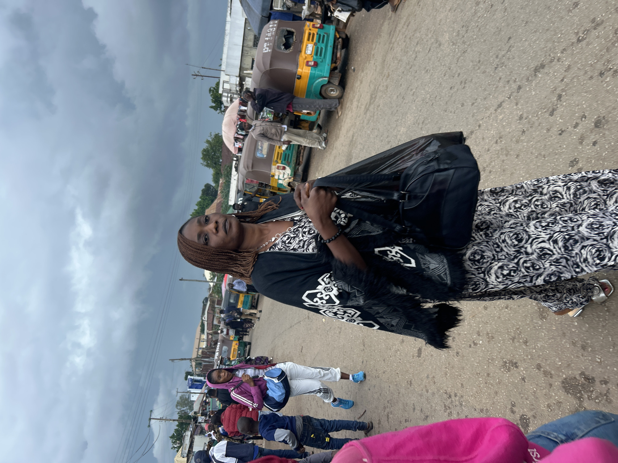 A medium shot captures a woman walking through a street scene in Jos, Nigeria, under an overcast sky. The woman, positioned in the foreground and slightly off-center to the right, wears a black and white patterned dress and a black shawl with a white pattern and fringed edges. She carries a black handbag in her right hand and a black plastic bag in her left. Her gaze is directed forward and slightly to the right.
Behind her, the street is populated with other individuals and several auto-rickshaws, known as "Keke Napep" in Nigeria. To her left, a young boy in a blue hoodie and jeans walks past, and further left, a woman in a purple hooded jacket with white stripes on the sleeves carries a blue and white duffel bag.
The environment appears to be a busy marketplace or transit hub. Buildings line the background, some with corrugated metal roofs, and overhead power lines are visible. The ground is a dusty, unpaved road. The auto-rickshaws, predominantly yellow and teal, are typical of local transportation in Jos. The overall atmosphere suggests a common, everyday scene in an urban setting in Nigeria.