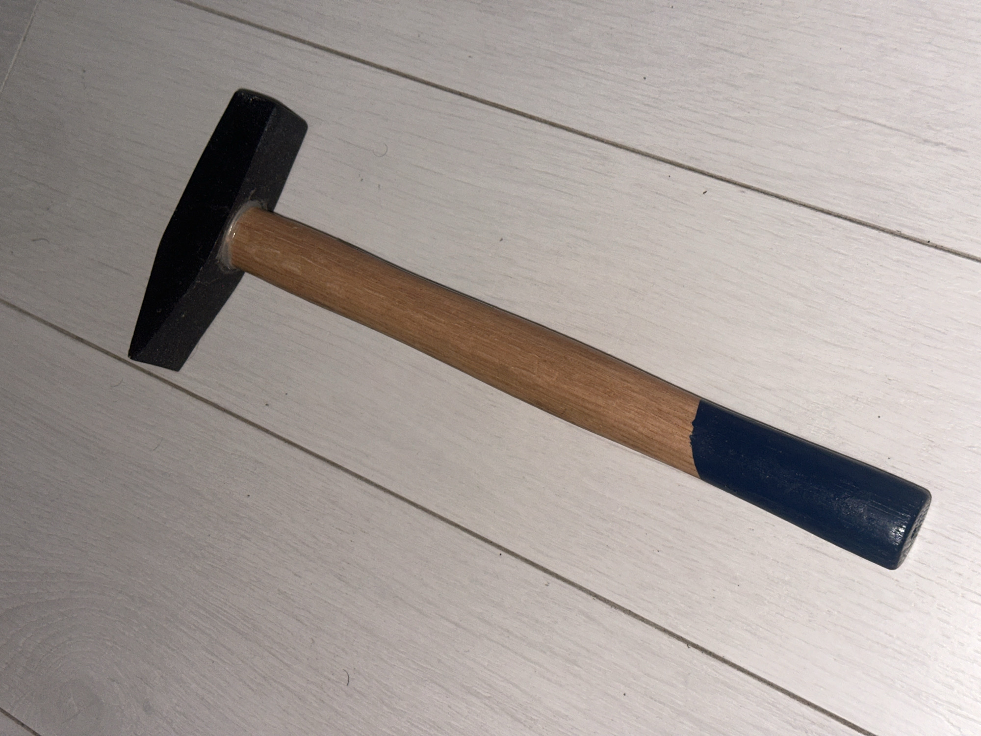A black-headed hammer with a wooden handle, painted dark blue at the grip, lies on a light-colored wooden floor with visible planks. The hammer is positioned diagonally across the frame. The scene is indoors and appears to be a residential setting. There is no indication of the location being the city of Sofia, Bulgaria. There are no people, actions, or interactions depicted.
