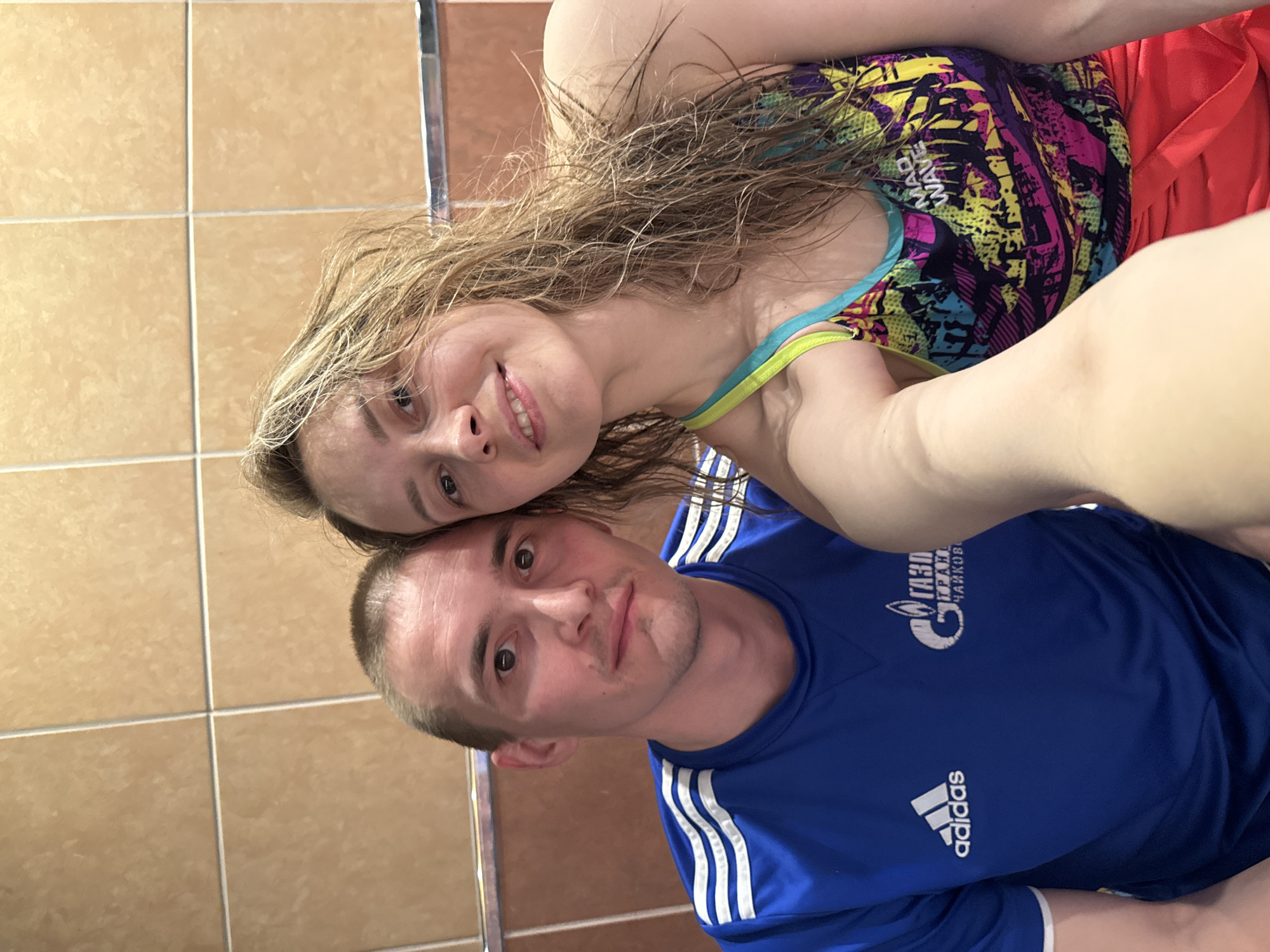 A selfie taken by a Caucasian male and female in what appears to be an indoor pool area with brown tile walls. The man is wearing a blue Adidas t-shirt with white stripes on the shoulders and the logo of the Russian company Gazprom (газпром) and the city name "Чайковский" printed on the upper right side of the chest, indicating a link to the city of Chaikovsky, Russia. The woman has wet, blonde hair and wears a colorful swimsuit. They are close together, looking into the camera. The lighting is bright, suggesting artificial illumination.