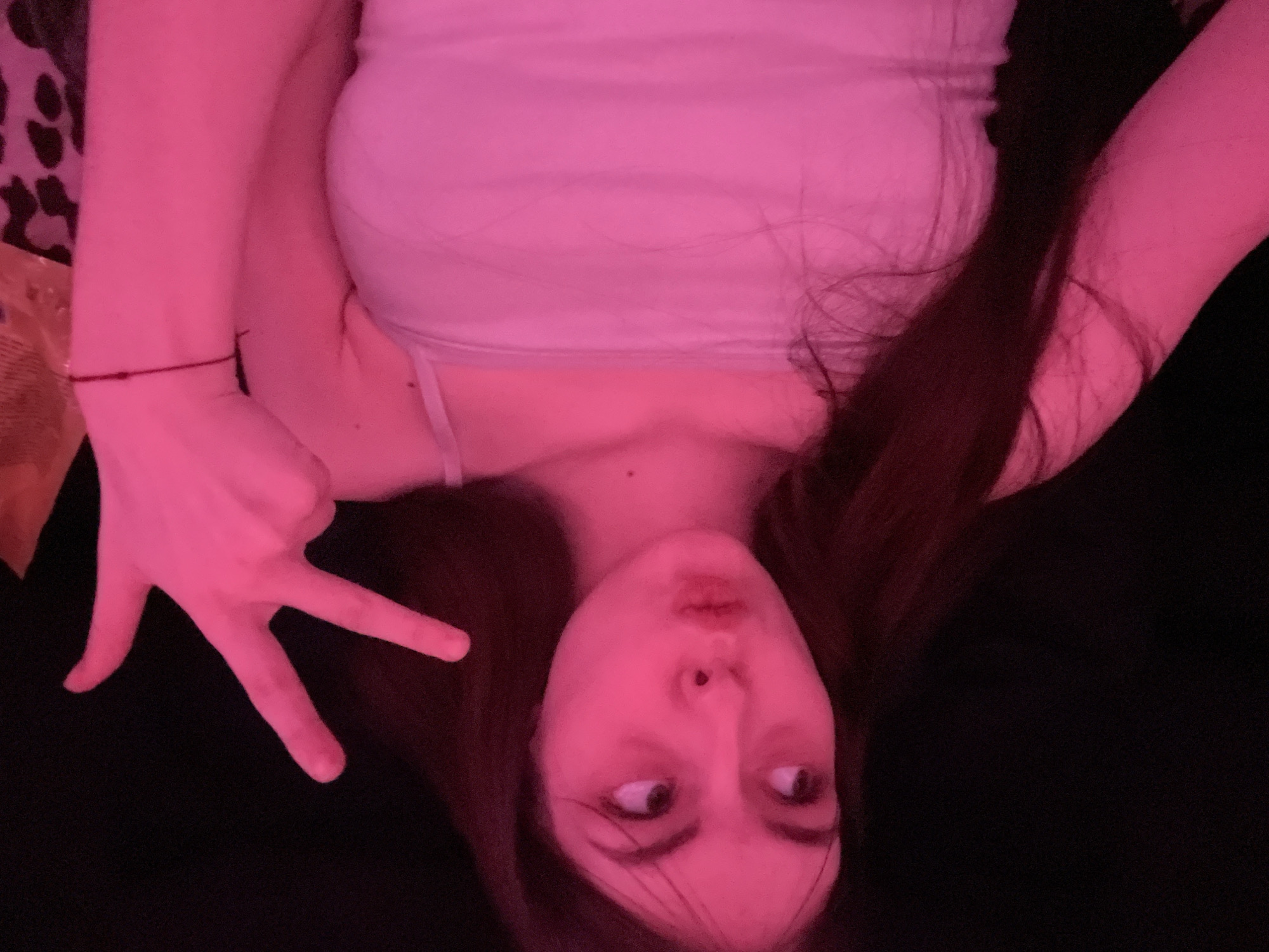 A person is shown in an upside-down, selfie-style photograph, posing with a hand raised in a gesture resembling a "W" or "3." The individual has fair skin, dark hair, and is wearing a light-colored tank top. The lighting is primarily pink, casting a strong hue across the subject and their surroundings. The background is predominantly dark, with a small patterned area visible on the left side, possibly indicating a piece of furniture or fabric. A thin, red bracelet is visible on the person's left wrist. No information suggests the image was taken in Saint Petersburg, Russia.