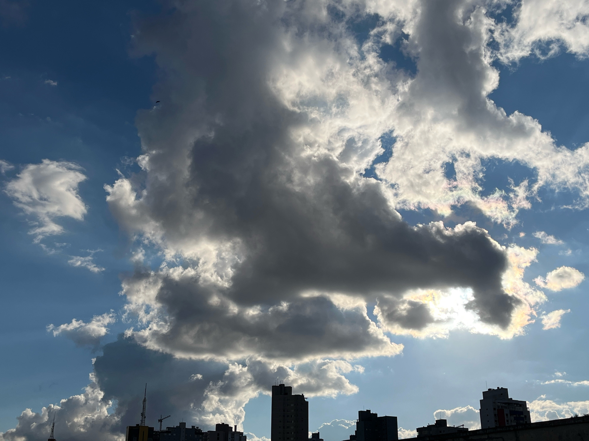 A photograph taken from a high vantage point in Cascavel, Brazil, captures a dramatic sky scene with towering storm clouds dominating the upper portion of the frame. The clouds are a mix of dark, ominous gray and bright, illuminated white, with the sun partially obscured behind them, creating a radiant glow and iridescent colors within the cloud formations. Below the clouds, a clear blue sky is visible in patches. Silhouetted against the sky are the rooftops of several urban buildings, their architectural details indistinct due to the backlighting. Some buildings appear to be high-rises, while others are shorter. The tops of at least two communication towers are also visible. The overall impression is one of an approaching or receding storm, with the city's skyline providing a grounding element to the dynamic sky.
