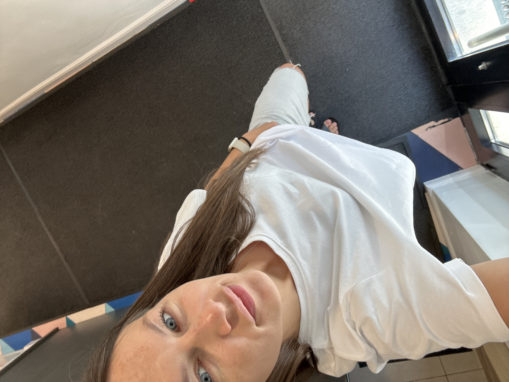 The image shows a medium-shot of a woman standing in what appears to be an indoor public space, possibly a retail store or lobby. She is wearing a white t-shirt and light-colored pants. The woman has long brown hair, blue eyes, and is looking at the camera with a slight smile. She is wearing a smartwatch on her left wrist and sandals on her feet. The floor is dark-colored carpet, and there are geometric patterns on the wall in the background, with colors like blue and pink. To the right, a reflective surface suggests a display case or window. The overall scene is well-lit and appears to be a candid or selfie-style photograph. There is no information to suggest this location is in Boreti, Montenegro.