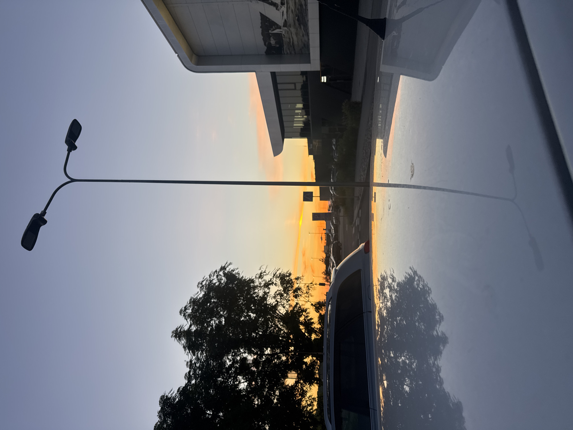 An eye-level, vertical, full outdoor shot captures a serene sunset scene at a parking lot in Tallinn, Estonia. The sky transitions from a pale blue to a vibrant orange, with the sun's rays illuminating the horizon. A tall street lamp with two lights stands prominently in the center, its dark silhouette contrasting with the colorful sky. To the left, silhouetted trees form a dense canopy, their branches reaching towards the sky. In the foreground, the reflective surface of a white car's roof mirrors the trees and the sunset, creating a striking visual effect. Further back, a modern building with a curved facade and large windows is visible on the right, its exterior adorned with a large black and white mural depicting sailboats. The parking lot is sparsely populated with a few cars. The overall atmosphere is tranquil and picturesque.