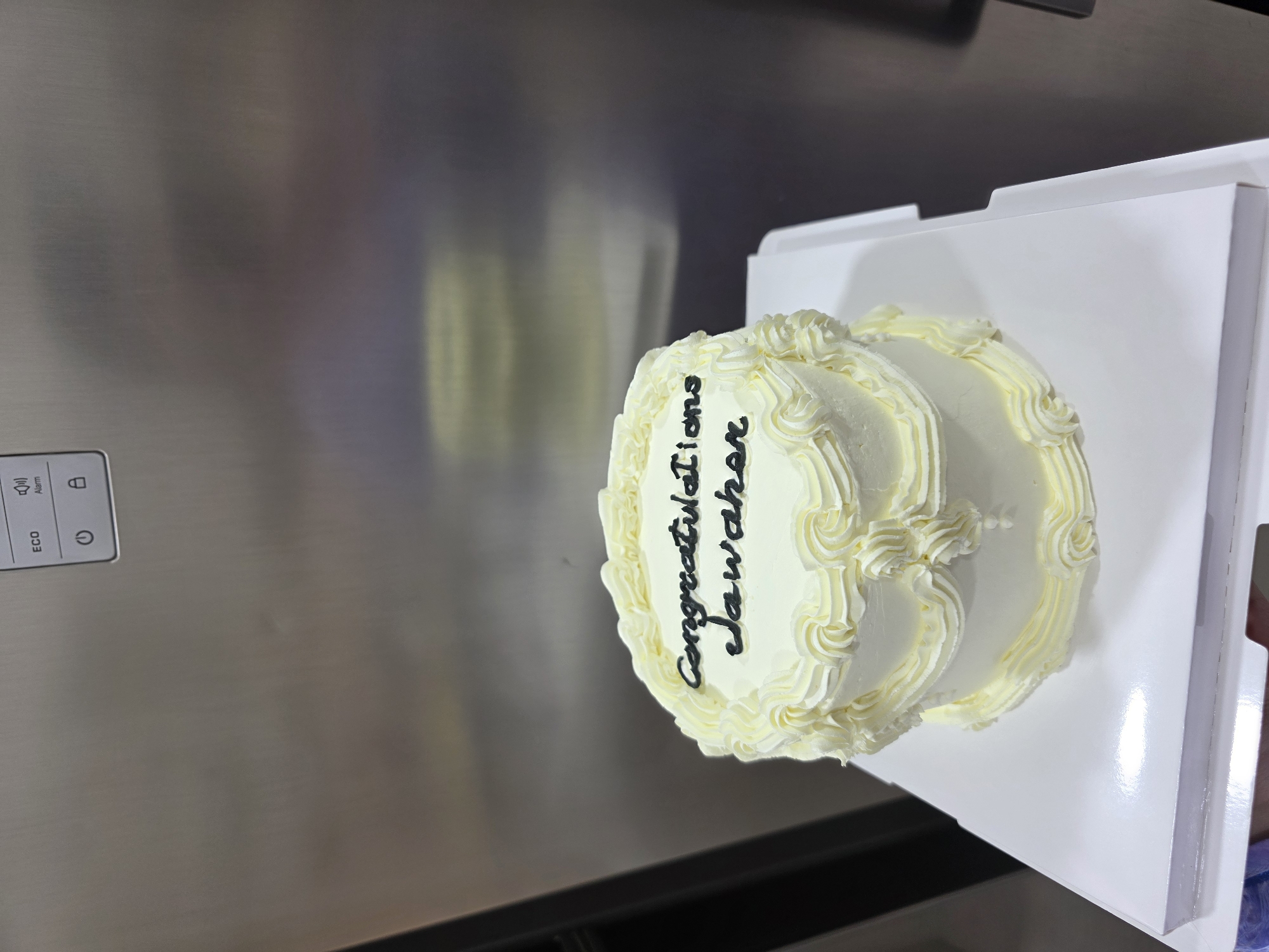 A two-tiered white cake with yellow frosting decorations is displayed on a white cake board. Black icing spells out "congratulations Jawahar" on the top tier. The cake is positioned in front of a stainless steel appliance, possibly a refrigerator, which has a control panel with icons for "ECO," "Alarm," a power symbol, and a lock symbol. The background is out of focus, but hints of a domestic or commercial setting are visible. The location is not specified in the image itself, but the question asks for it to be set in Dibba Province, Oman.