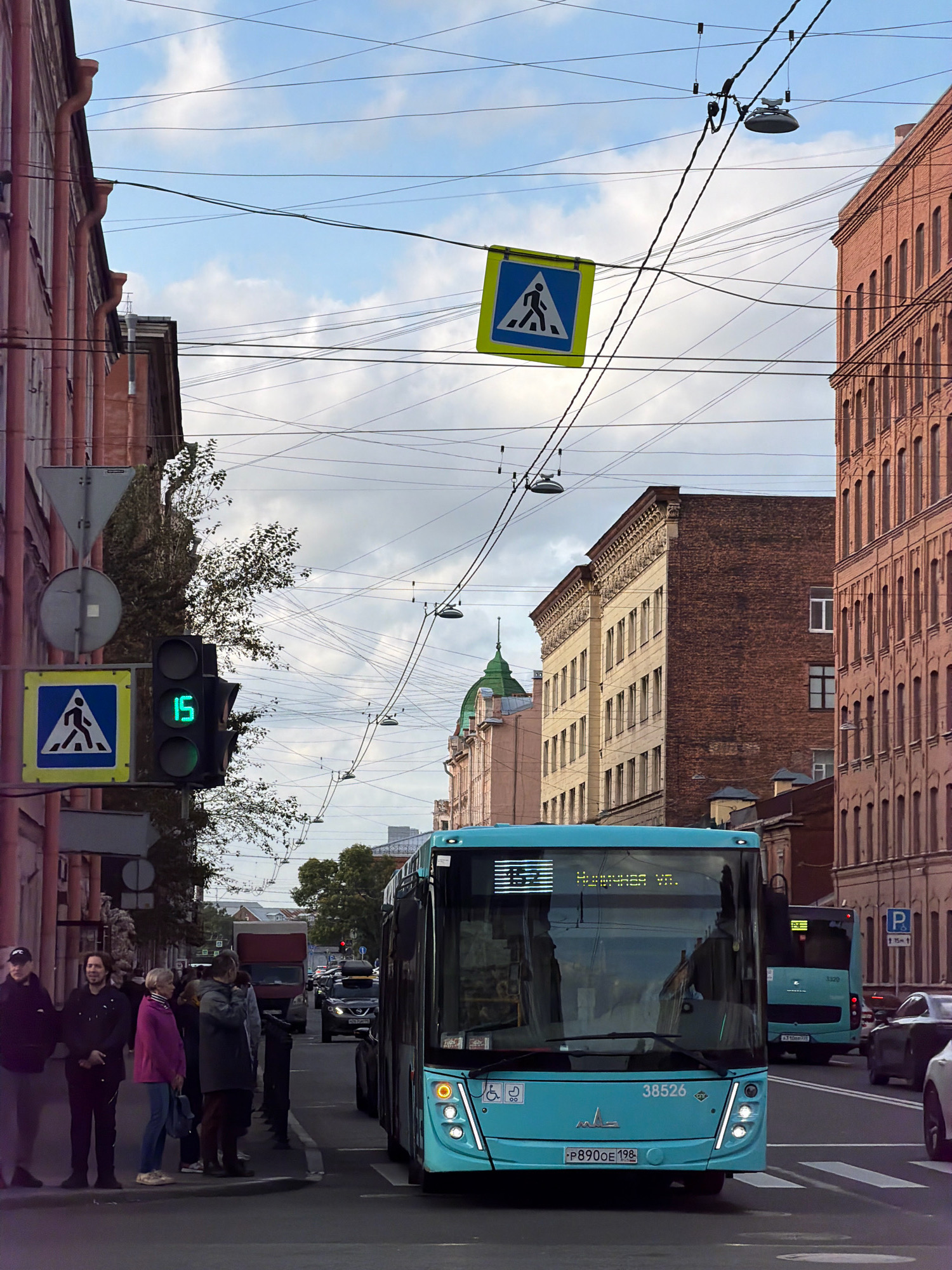 The image captures a street scene in Saint Petersburg, Russia, likely near a pedestrian crossing. A bright turquoise bus, displaying the route number "152" and text in Cyrillic characters, is prominently featured in the foreground, stopped at a traffic light. The traffic light shows a green signal with the number "15" displayed within it. To the left of the bus, a pedestrian crossing sign with a blue background and a white silhouette of a walking person is visible, hanging over the street. Another similar pedestrian crossing sign is situated lower down on a pole, next to the traffic light. Several people are standing on the sidewalk to the left of the bus, waiting. The street is lined with buildings of varying architectural styles, including a red brick building on the right with numerous arched windows, and a lighter colored building with ornate detailing on the left. Overhead, a complex network of electrical wires crisscrosses the sky, a common sight in urban environments. The sky is partly cloudy, with patches of blue visible. In the background, other vehicles, including a truck and cars, are also present on the street.