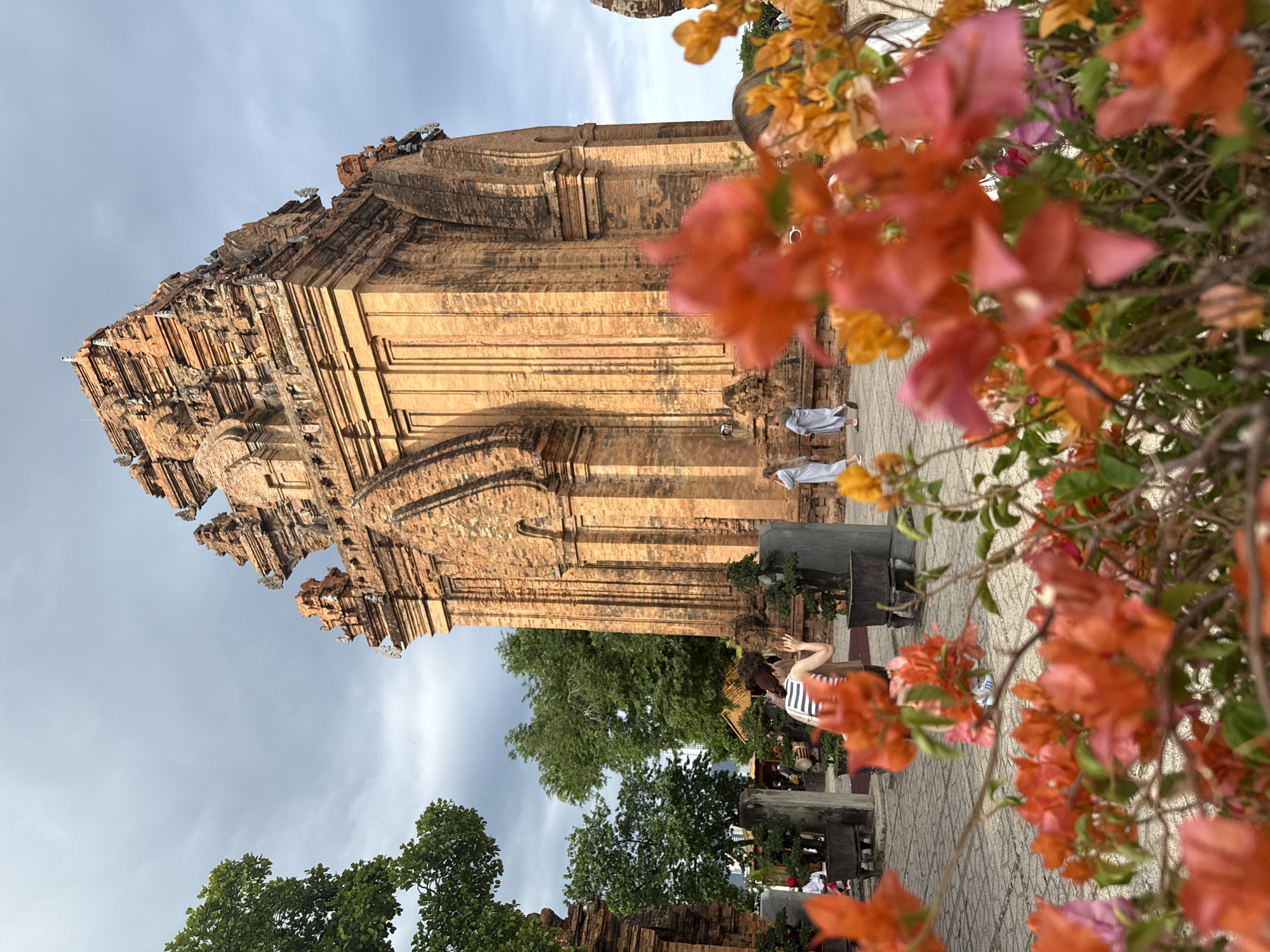 This image captures the Po Klong Garai Cham Towers, a historical site located in the city of Nha Trang, Vietnam. The main subject is a large, ancient brick tower with intricate architectural details, typical of Cham craftsmanship. In the foreground, a cluster of vibrant pink and orange bougainvillea flowers are in soft focus, framing the scene.
Several people are visible in the mid-ground. To the left, a woman wearing a striped tank top and shorts is standing and looking towards the tower, with her hand raised as if taking a picture or waving. She is positioned near some bonsai trees in planters. Further to the right, two individuals in light-colored robes, possibly monks or tourists in traditional attire, are walking away from the camera. Another person, partially obscured by flowers, is on the far right.
The ground is paved with stone tiles, and there are more bonsai trees and stone elements scattered around the courtyard. The sky above is overcast with clouds, suggesting a temperate day. The overall atmosphere is one of historical exploration and natural beauty, with the ancient architecture juxtaposed against the lush flora and the presence of visitors.