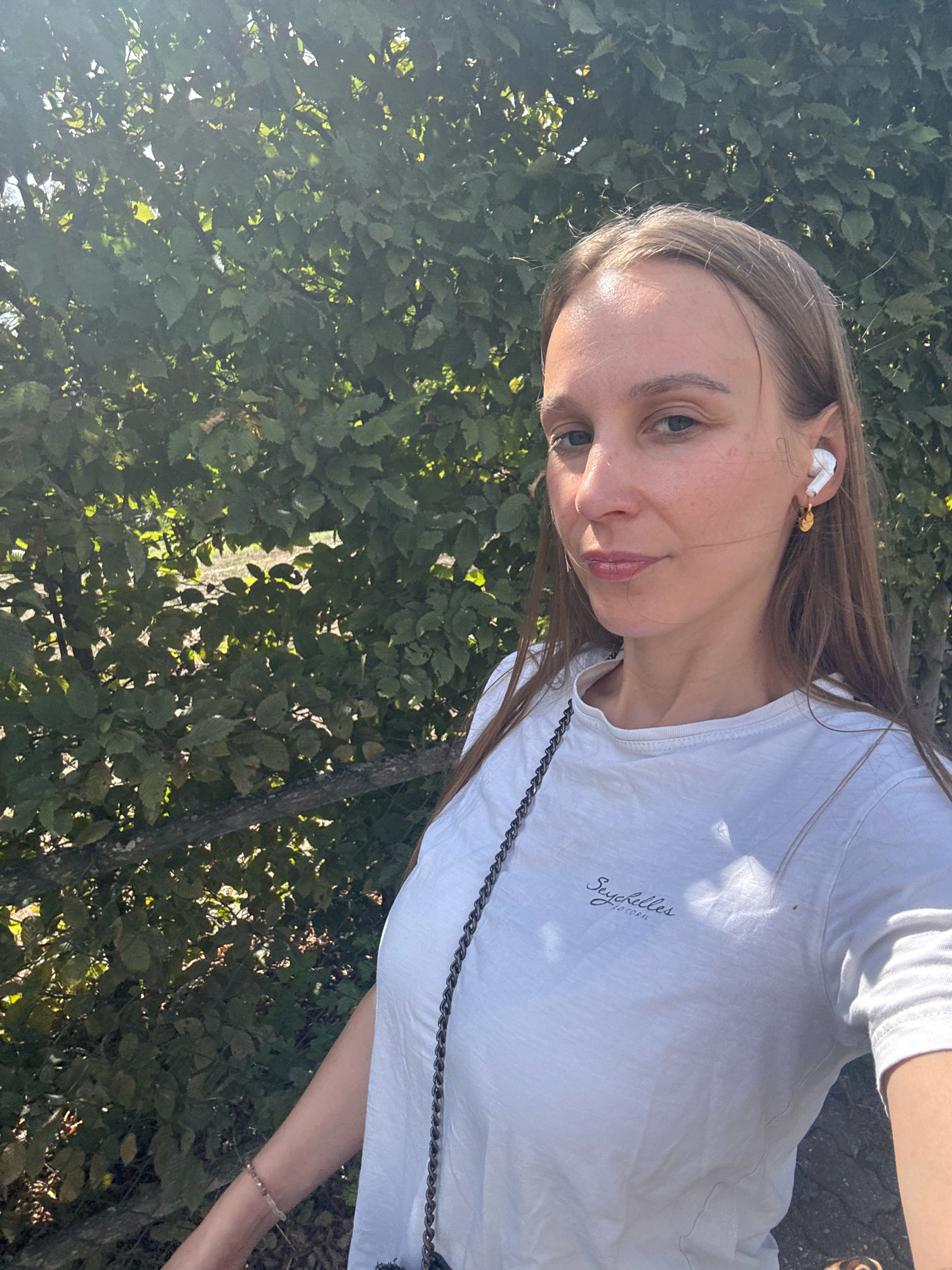 A young woman with long, light brown hair, wearing a white t-shirt and wireless earbuds, takes a selfie outdoors. The background is filled with lush green foliage, suggesting a park or natural setting. A portion of a wooden fence is visible in the lower left of the image. The woman is looking directly at the camera with a slight smile. The lighting indicates it is daytime, with sunlight dappling through the leaves. The image does not provide any specific clues about the location within Überherrn, Germany.
