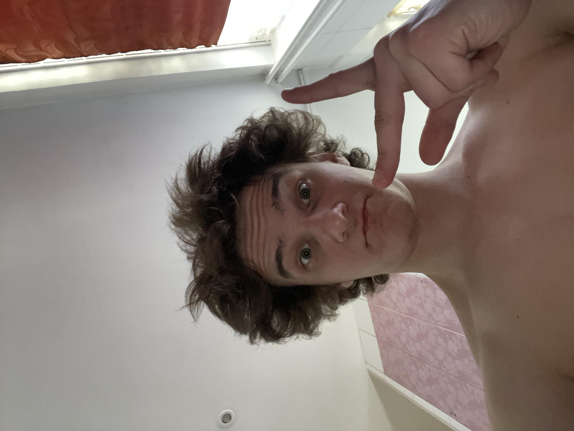 A shirtless individual with curly brown hair is taking a selfie in a bathroom. The person is looking at the camera with a neutral expression and making a hand gesture. The background shows white walls, a pink-tiled wall, and a shower curtain. There is no specific indication of this image being taken in Krasnoyarsk, Russia.