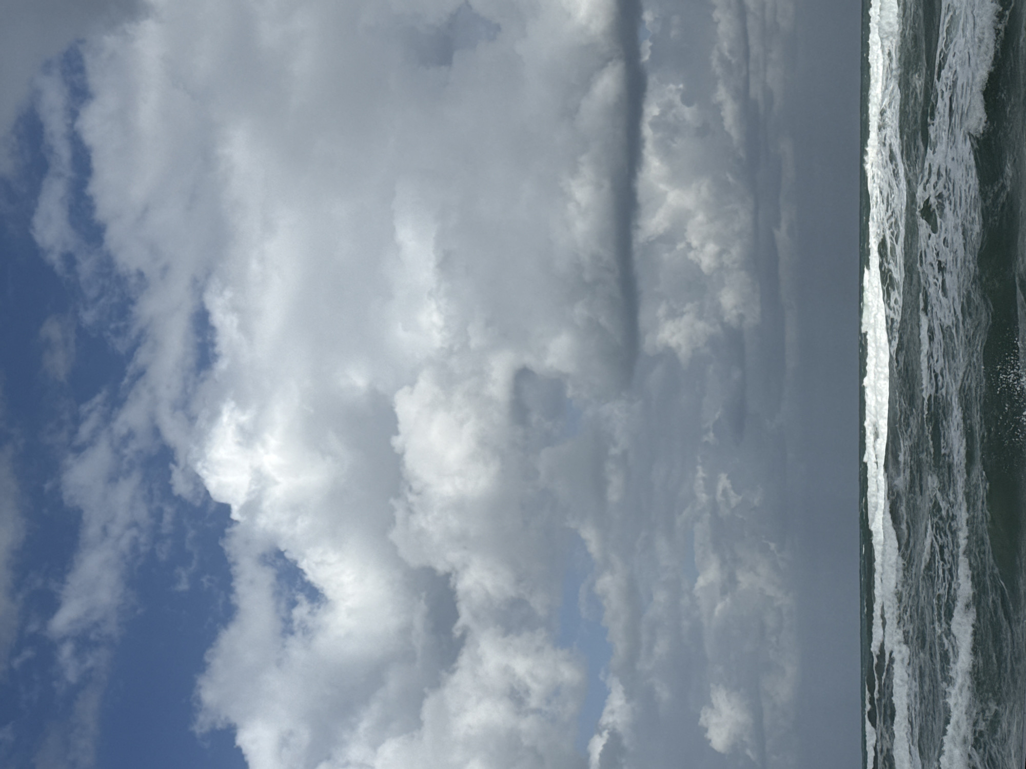 The image depicts a scene at the beach, likely in Tel-Aviv, Israel. The sky is filled with various cloud formations, with a patch of blue visible towards the left. The ocean is visible on the right side of the frame, with breaking waves and foamy water. The horizon line is faint. There are no people visible in the image.