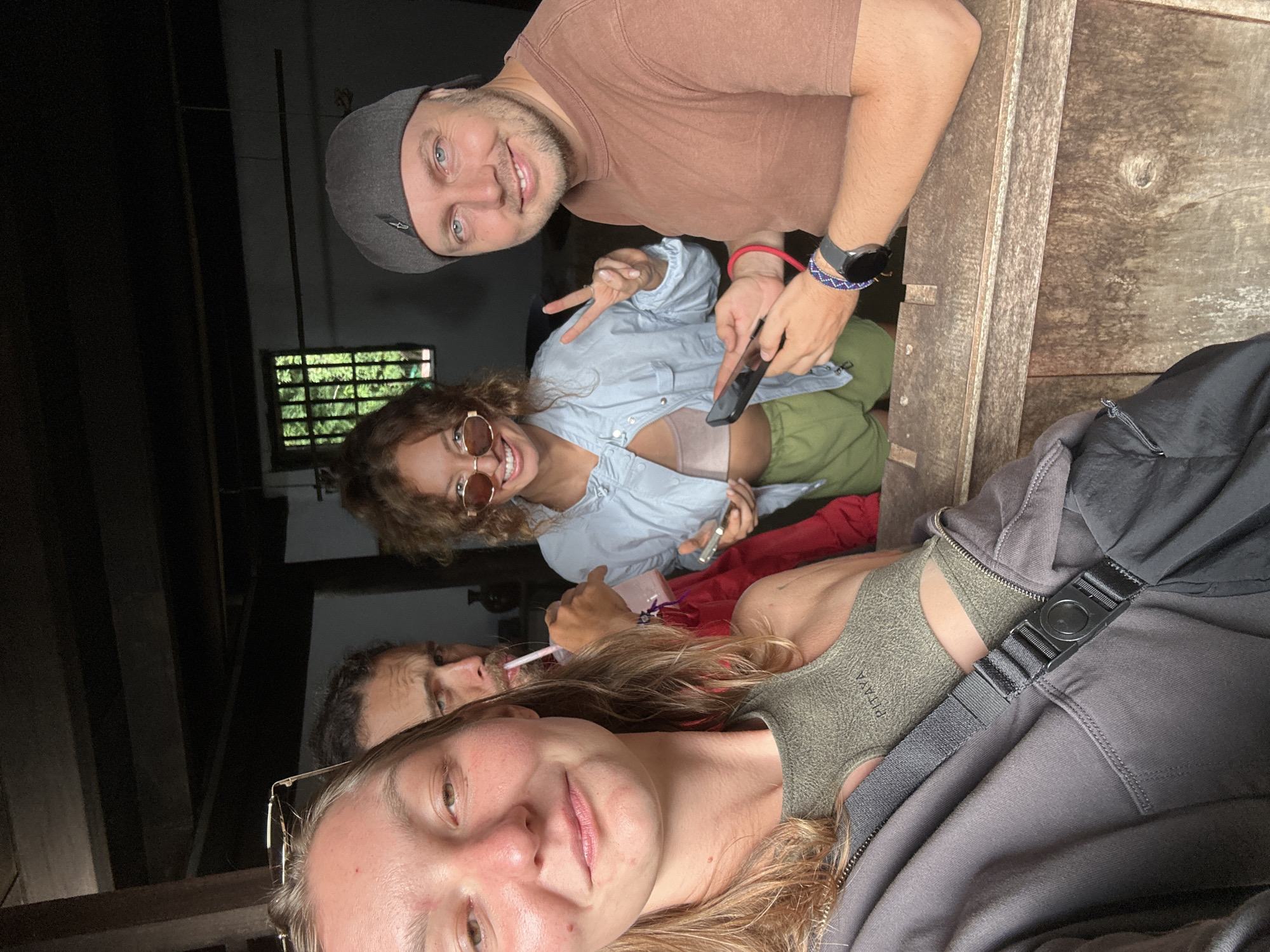 In Sà Phìn Commune, Vietnam, a group of four people are in what appears to be a rustic, dimly lit interior, possibly a wooden structure or an old building. The photo is taken from a low angle, focusing on the faces of the individuals. The person closest to the camera, likely taking the selfie, is a woman with light hair and eyeglasses. Behind her is another person drinking from a cup with a straw. Further back, a woman with curly hair and sunglasses is making a peace sign while holding a phone. A man in a cap and a brown shirt is looking at the camera with a smile. The environment suggests an informal gathering or a stop during travel. There's a wooden paneling visible in the background. The lighting is dim, emphasizing the interior setting.