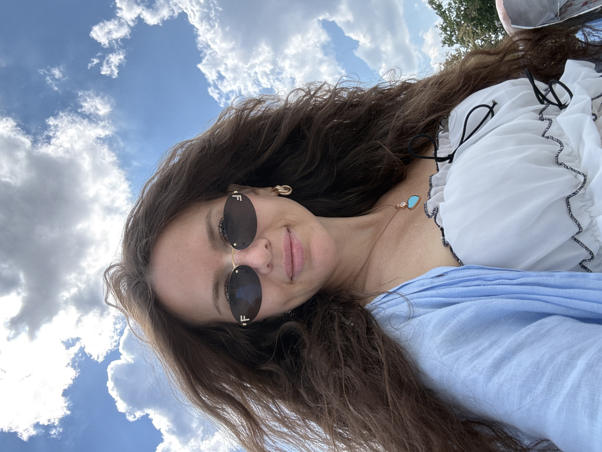 A woman with long, brown, curly hair is taking a selfie outdoors. She is wearing oval-shaped sunglasses with gold frames and an "F" logo on them. She also has on gold hoop earrings, a gold necklace with a turquoise pendant, and a light blue open cardigan over a white ruffled top with black trim. The background consists of a bright blue sky with fluffy white clouds and some greenery visible on the right side of the image. The lighting suggests it is daytime.