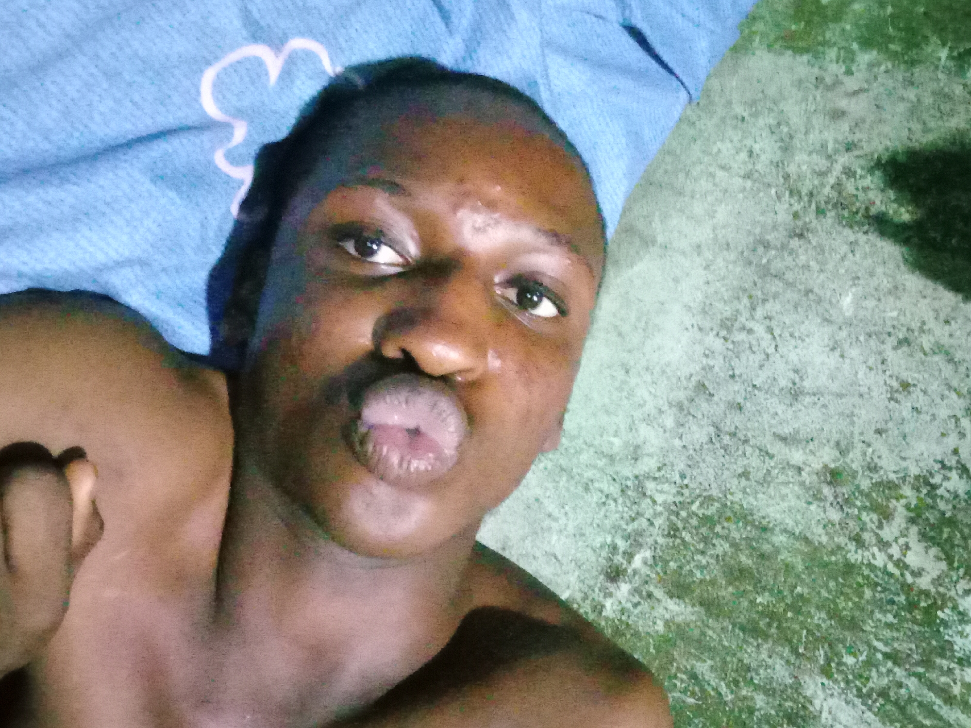 The image, taken in Nkpor, Nigeria, is a close-up, low-angle, overhead shot of a person lying on a light blue, textured surface with a white floral pattern. The background is a rough, mottled gray and green surface. The person has dark skin and is shirtless. Their head is tilted back, and their eyes are open. Their lips are puckered, forming a kiss-like expression. A hand is visible, resting near the person's chest. The lighting is dim, casting shadows and highlighting the textures of the surfaces and skin.