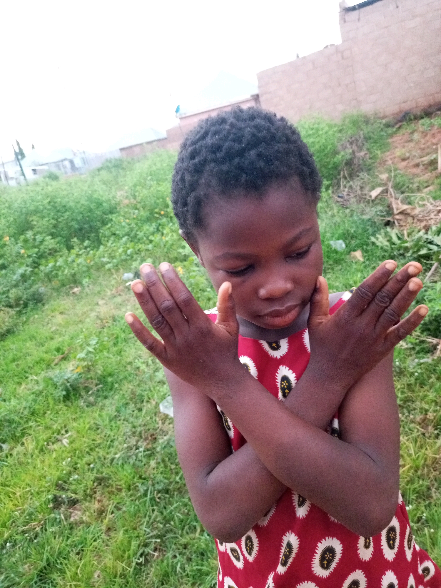 A young girl with dark skin and short, black, curly hair is standing in an outdoor environment. She is wearing a red dress with a pattern of white ovals containing black and yellow elements. Her arms are crossed in front of her chest, and her hands are positioned with her palms facing outwards. She is looking downwards. The background consists of green grass, some dry vegetation, and in the distance, buildings with brick walls and roofs are visible. The sky is overcast and appears bright. The overall setting suggests a rural or semi-urban area, possibly within Tempure, Nigeria.