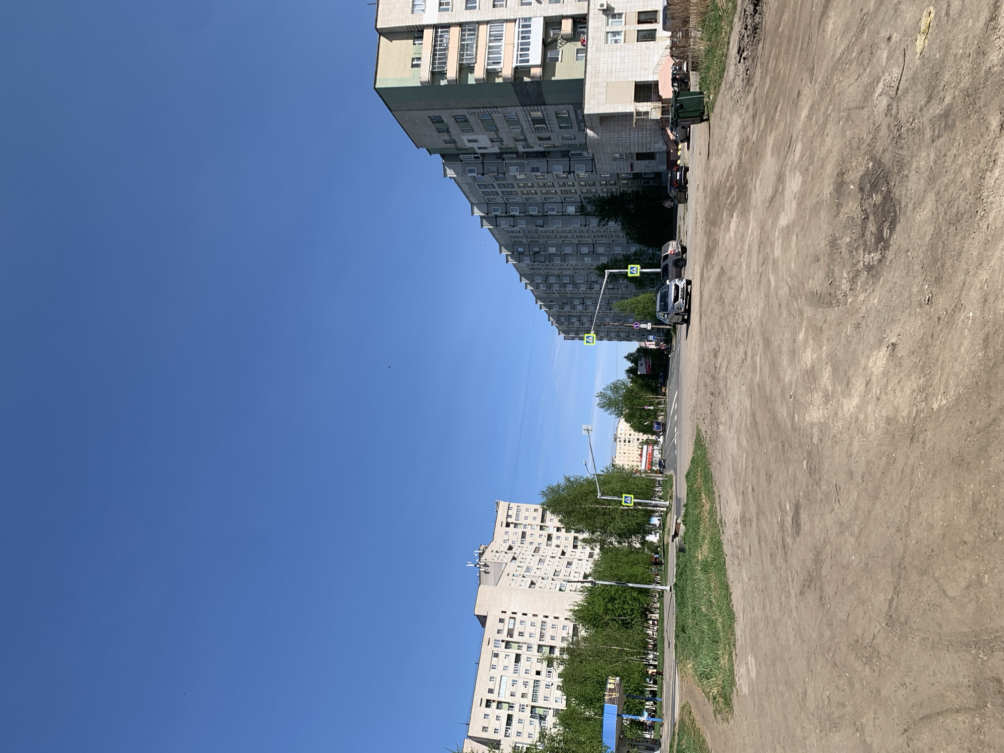 The image depicts a street scene in what appears to be a residential area in Severodvinsk, Russia. The perspective is oblique. On either side of the street, tall apartment buildings with repetitive facade designs typical of Soviet-era architecture are visible. The sky is clear and blue. The street shows signs of wear and includes pedestrian crosswalks with signage. There are cars parked along the side of the street and trees lining the road. The ground adjacent to the street is dirt. No people are clearly visible.