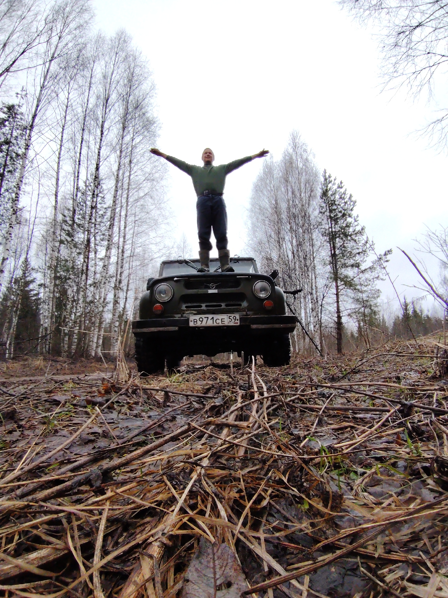 A low-angle outdoor shot captures a man standing triumphantly on the hood of a dark green, rugged off-road vehicle in a sparse forest. The man, fair-skinned and wearing a dark green long-sleeved shirt, dark pants, and boots, has his arms outstretched horizontally, gazing upwards or straight ahead, conveying a sense of freedom or achievement.
The vehicle, appearing to be a Soviet-era UAZ-469 or similar 4x4, is positioned on a muddy, debris-strewn forest floor, covered thickly with fallen dry leaves, twigs, and branches. Patches of green grass are visible amidst the brown and wet ground, indicating damp conditions. The forest in the background consists primarily of tall, slender birch trees with white bark, mostly bare of leaves, interspersed with some darker evergreen trees. The sky is overcast and bright, suggesting daytime with diffused light.
A Russian license plate, "B971CE 59 RUS," is clearly visible on the front of the vehicle. The overall scene depicts an off-road adventure or exploration in a remote, natural environment, consistent with a wilderness area in Markovskii, Russia.