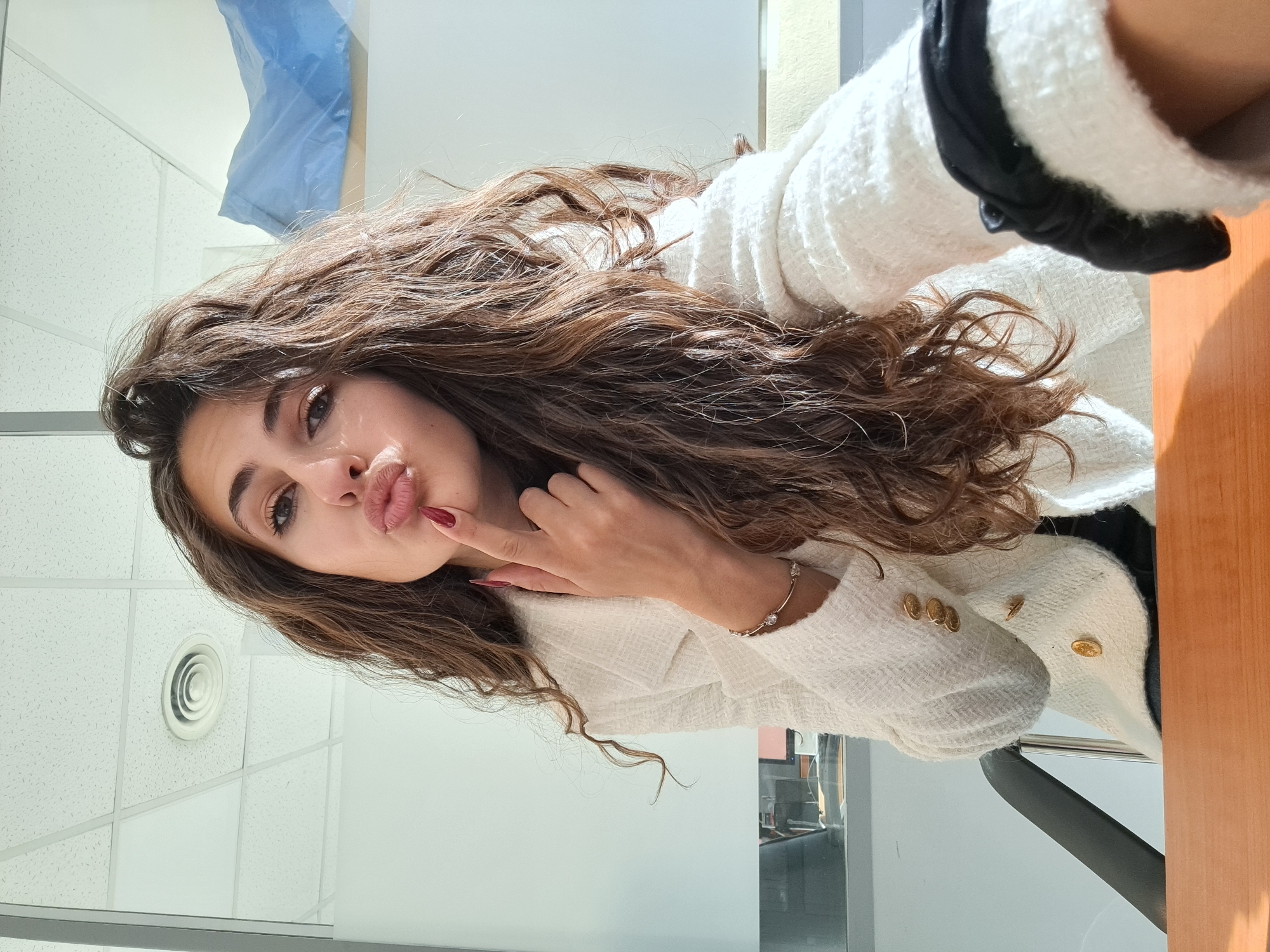 A woman is taking a selfie indoors. She has long, curly brown hair and is wearing a white textured blazer with gold buttons over a black top. She is puckering her lips and holding a finger to her cheek. Her left hand has red nail polish. She is wearing a bracelet on her right wrist. The background includes a white ceiling with square panels and a circular air vent, a light blue object hanging on the wall, a wooden surface, and a black chair. The location is indoors, but the precise location in the city of Sofia, Bulgaria, cannot be determined from the given image.