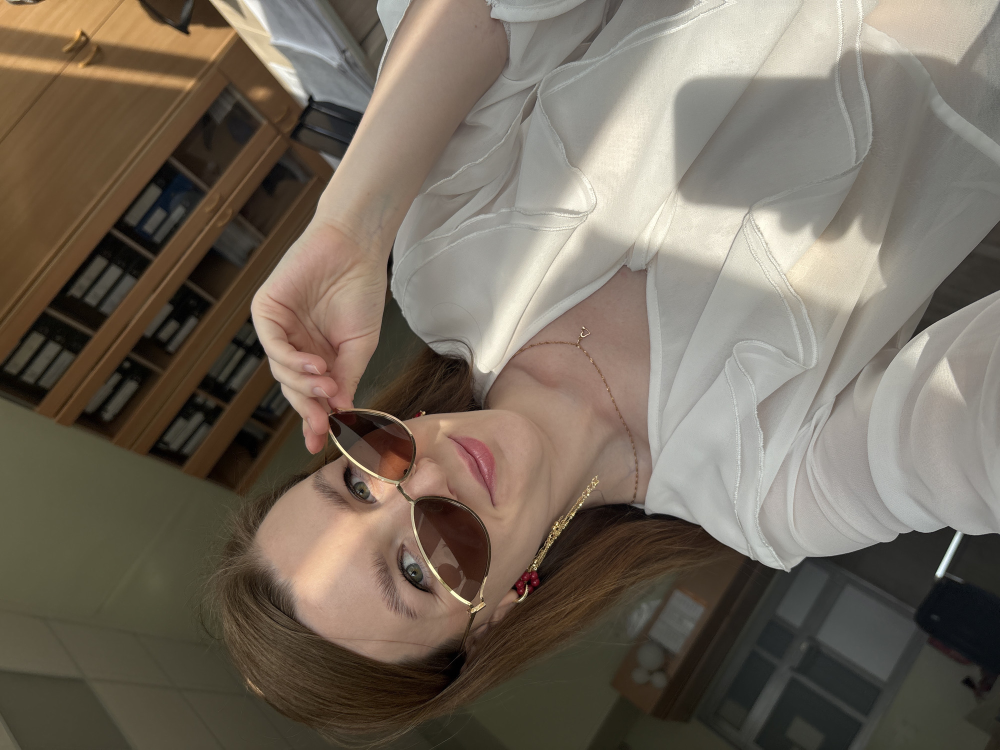 A young woman is seen in a selfie, holding sunglasses in her hand. She has light skin, green eyes, and brown hair. She wears a white blouse and a gold necklace. A gold earring is visible on her left ear. The background shows a cabinet and a window. There is no explicit indication that the location is Izhevsk, Russia, in the picture.