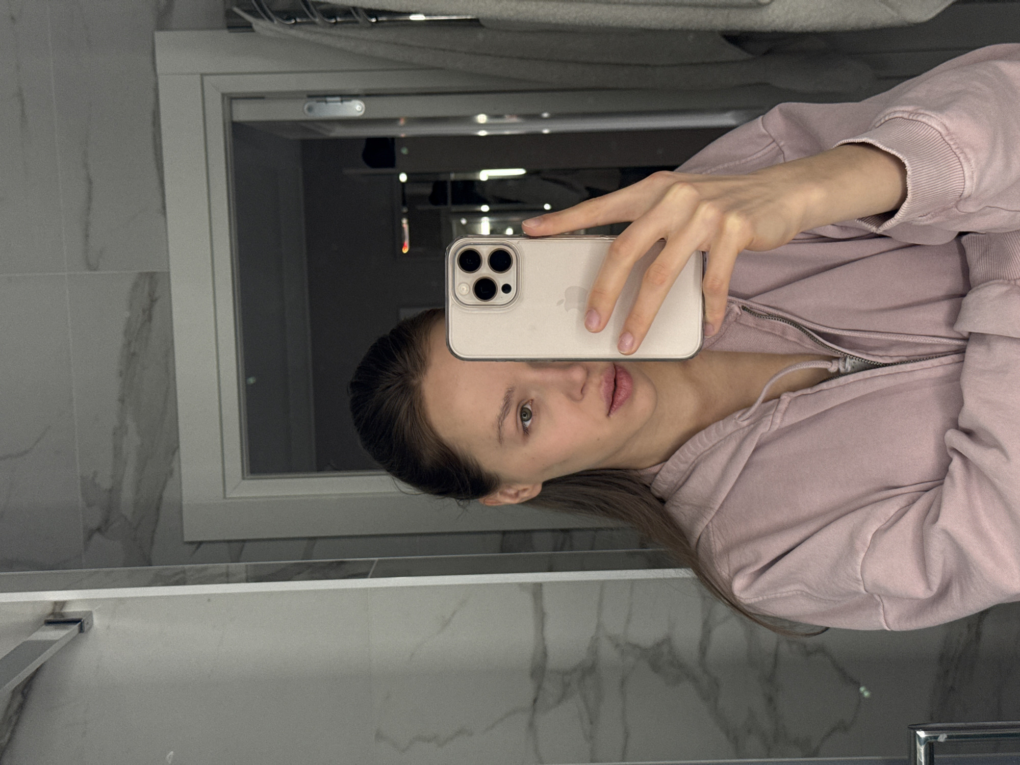 The image shows a young woman taking a mirror selfie in a bathroom. She is seen from the chest up, positioned to the left, holding a light-colored iPhone (likely a Pro Max model with its distinct three-camera array) in her right hand. The phone is encased in a clear protective cover, and the Apple logo is visible on its back.
The woman wears a light pink or mauve zippered hoodie. Her long, dark brown hair is partially pulled back, with some strands framing her face. She has a neutral expression, looking directly into the camera's reflection, with her lips slightly parted.
The background reflected in the mirror reveals elements of a modern bathroom. White marble-patterned tiles adorn the walls. A white bathtub or large sink with a silver faucet is visible in the lower portion of the reflection. A white door or window frame can also be seen. The lighting suggests an indoor setting, likely under artificial illumination. No text is visible in the image.