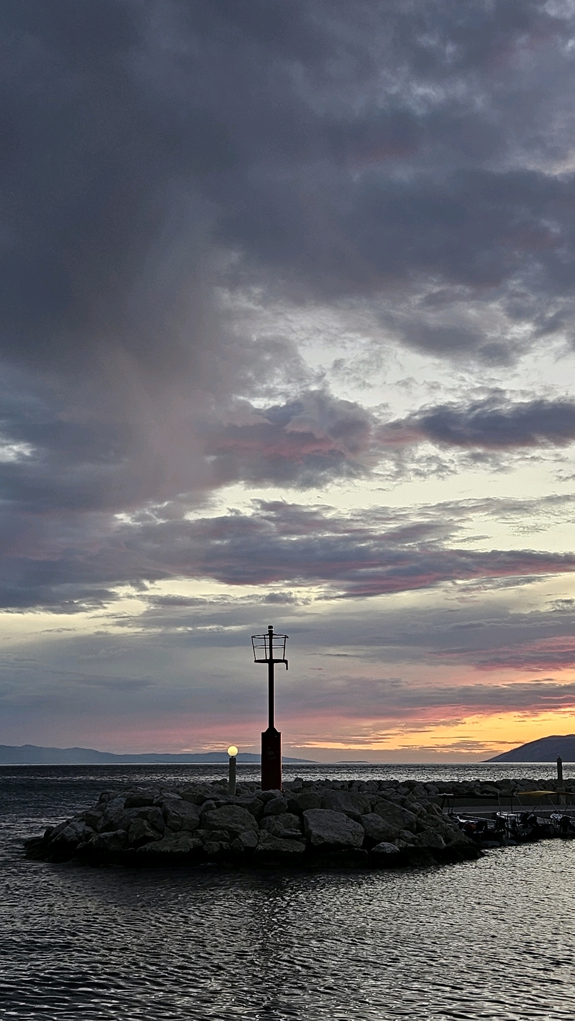 The image depicts a twilight scene on the Adriatic coast in Općina Tučepi, Croatia. In the foreground, dark, rippling water fills the lower portion of the frame. A rocky jetty extends into the water, topped with a red and black lighthouse or marker and a small, illuminated post. Beyond the jetty, a calm sea stretches to the horizon, where distant hills or islands are silhouetted against a sky ablaze with the warm colors of sunset, transitioning from oranges and yellows near the horizon to purples and grays overhead, punctuated by dramatic clouds. A few small boats are moored to the right of the jetty. The overall atmosphere is serene and evocative of the end of the day.