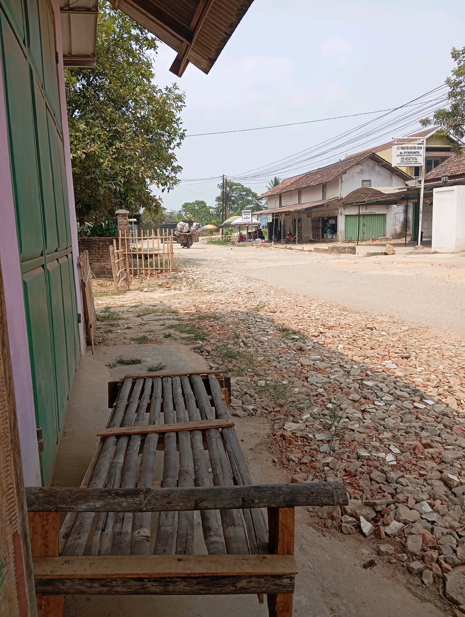 The image depicts a street scene in what appears to be a rural or semi-urban area, possibly in the city of Batang Hari Ogan, Indonesia. In the foreground, on the left, is a section of a building with a pale pink and green facade, partially obscuring the view. A simple, rustic bench made of bamboo and wood sits in front of this building, facing a sandy path.
The path leads to a dirt road where several structures are visible along the right side. These buildings appear to be shops or small businesses, some with open storefronts and others with closed, corrugated metal or green doors. A sign for "PRAKTEK DOKTER UMUM dr. FITRYANTO" is prominently displayed on one of the buildings. There are utility wires strung across the sky.
On the road, a motorcycle is parked in front of one of the shops. Further down the road, a person is seen riding a motorcycle laden with goods, heading away from the camera. Colorful umbrellas are visible further down, suggesting the presence of stalls or outdoor vendors. The ground along the road is covered with loose stones and debris, indicating a potentially unpaved or under-construction area. Trees and vegetation are present in the background, and the sky is overcast. The overall impression is one of a quiet, working-class street in a developing area.
