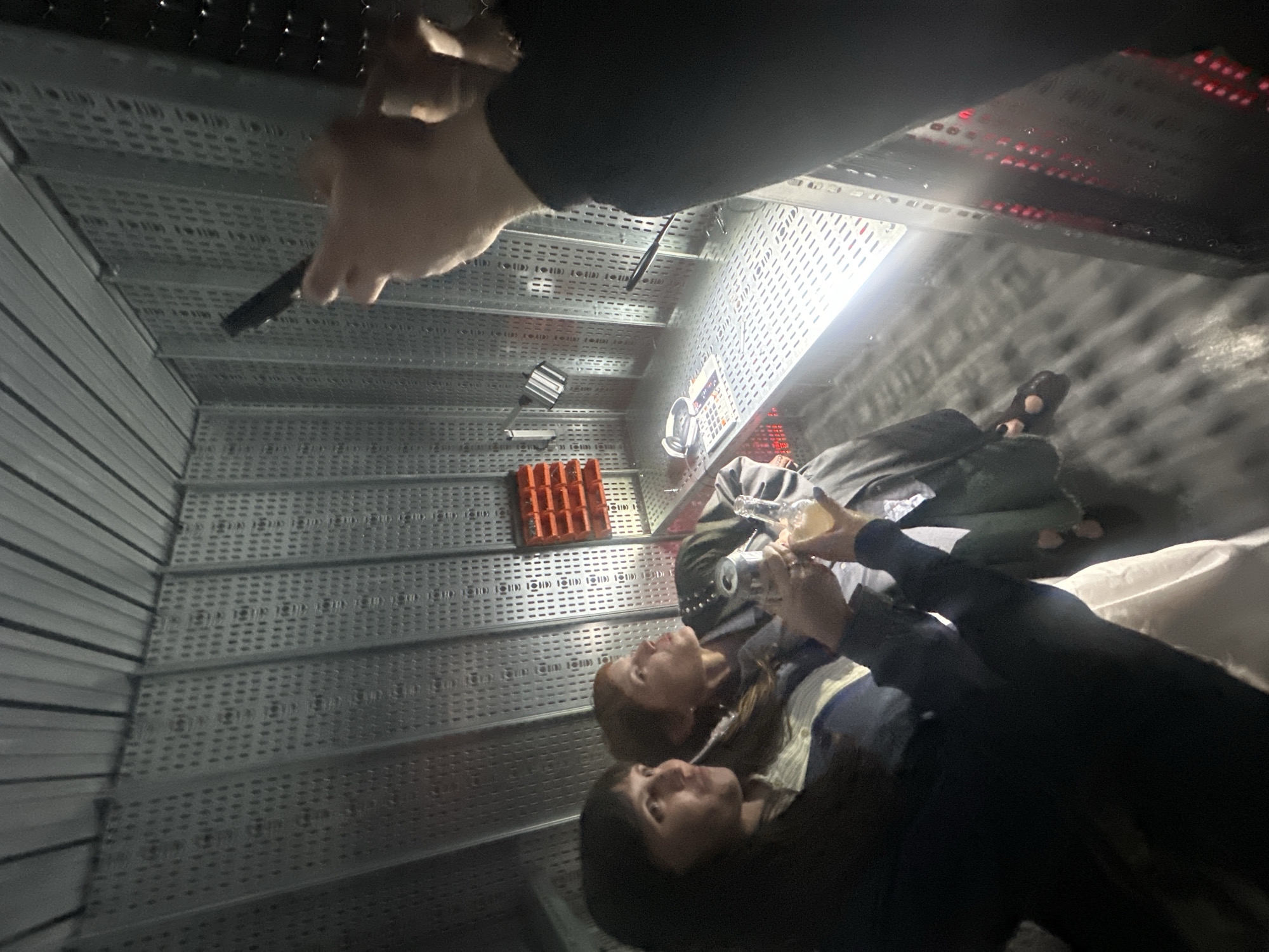 This media file captures an indoor scene with a distorted, high-angle perspective, giving the impression of looking down into a confined, industrial-style space, possibly an escape room or a themed venue in London, United Kingdom.
The setting is characterized by walls and surfaces made of metallic, perforated grating panels, suggesting a utility shaft, server room, or a futuristic environment. Bright artificial lights illuminate the scene, creating strong reflections on the metal and casting shadows. In the far background, areas of red light, possibly from electronic equipment or indicators, are visible.
Several people are present. In the foreground, a hand, likely belonging to the person taking the photo, is seen holding a small black object, resembling a pen or a thin flashlight. Further into the frame, two women are partially visible, looking upwards or towards each other. One woman has reddish-brown hair and is wearing a jacket, while the other, with dark hair, wears a striped shirt. Their hands are clasped around a clear glass bottle, suggesting they are sharing a drink, with a silver can also nearby. Behind them, another person is lying down, with their feet visible, adding to the unusual composition.
Other objects include an orange, grid-like tray or box structure resting on one of the metallic shelves or panels. In the background, what appears to be a control panel or electronic device with several red illuminated buttons or lights is visible, along with white headphones.
The activity appears to be social interaction within an unusual and perhaps challenging environment, consistent with an entertainment experience. There is no clearly visible text in the image. The artificial lighting and lack of exterior views suggest it is either night or a windowless indoor setting.