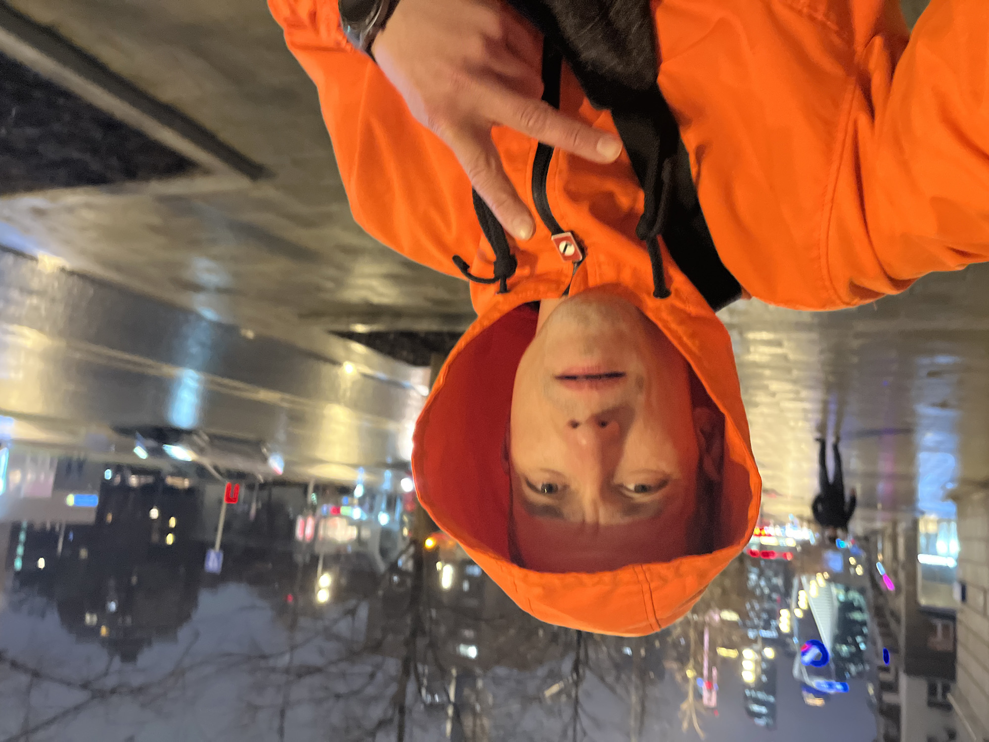 A person is taking a selfie at night on a wet street in Moscow, Russia. The person is wearing an orange hoodie, a watch on their wrist, and making a "peace" sign with their fingers. The background shows a cityscape with streetlights and illuminated buildings reflected on the wet pavement. Another person is walking away in the background. Trees without leaves are also visible.