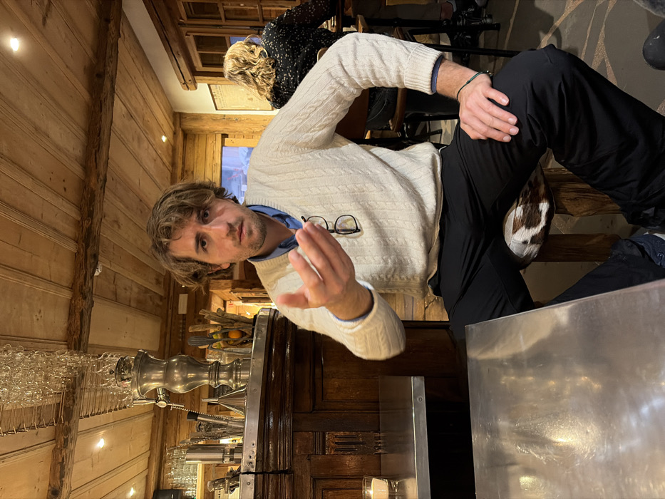 This image displays an interior scene within an establishment in La Clusaz, France, characterized by a rustic, chalet-style decor with extensive use of wood.
A male individual, appearing to be in his late 20s to early 40s, is seated in the foreground, facing the viewer. He has medium-length, wavy light brown hair. He is wearing a cream-colored cable-knit V-neck sweater over a light blue collared shirt and black trousers. Prescription glasses are hooked onto the V-neck of his sweater. He is seated on a wooden stool featuring a brown and white cowhide-patterned seat. His left hand is extended forward, palm slightly open with fingers partially curled, in a gesturing position. His right hand rests on his right thigh, with a thin green bracelet visible on his wrist. His expression is neutral, and he maintains direct eye contact with the camera. A reflective stainless steel tabletop is visible in the immediate foreground to the left.
To the left of the male subject, a dark wooden bar counter with ornate carvings is visible. Above the counter, an array of clear drinking glasses hangs upside down from a metal rack. A tall, ornate silver-colored tap or dispenser stands prominently on the bar, flanked by various bar tools and bottles. A small, lit candle in a clear glass holder is present on the stainless steel table to the left.
In the mid-ground to the right, a second individual, a female with wavy blonde hair, is seated facing away from the camera, wearing a dark top with a light patterned design. The overall environment features light-colored wooden paneled walls and a wooden beamed ceiling, incorporating recessed lighting fixtures. A window or doorway is partially visible in the distant background, allowing for a glimpse of an outdoor setting.